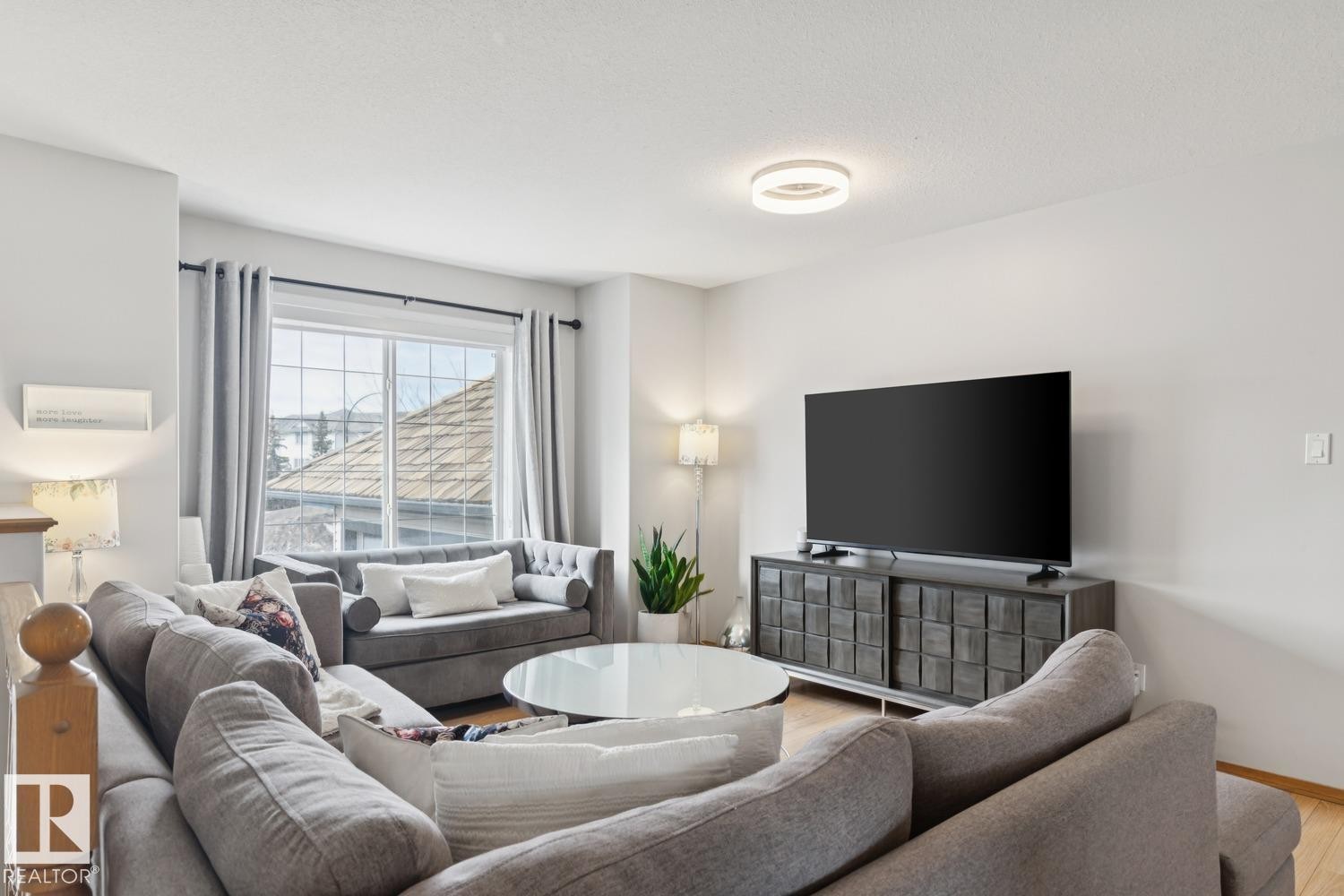 Spacious living area featuring light wood-finish flooring, a contemporary ceiling light fixture, large window with curtains, and neutral wall tones - 1 225 Blackburn Drive E, Edmonton, AB