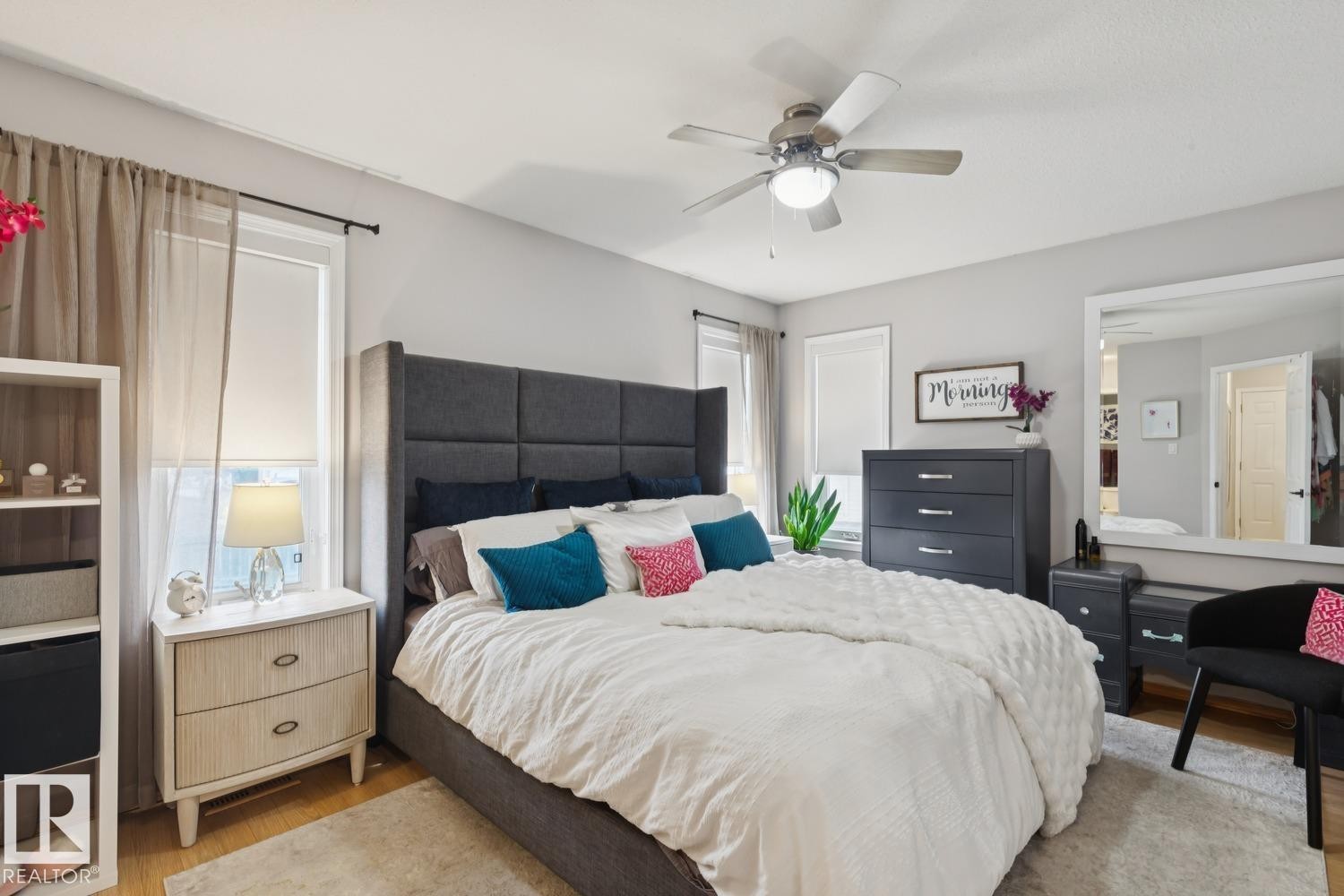 Spacious bedroom featuring light wood-finish flooring, a ceiling fan with integrated lighting, multiple windows with roller shades, and light gray wall tones - 1 225 Blackburn Drive E, Edmonton, AB