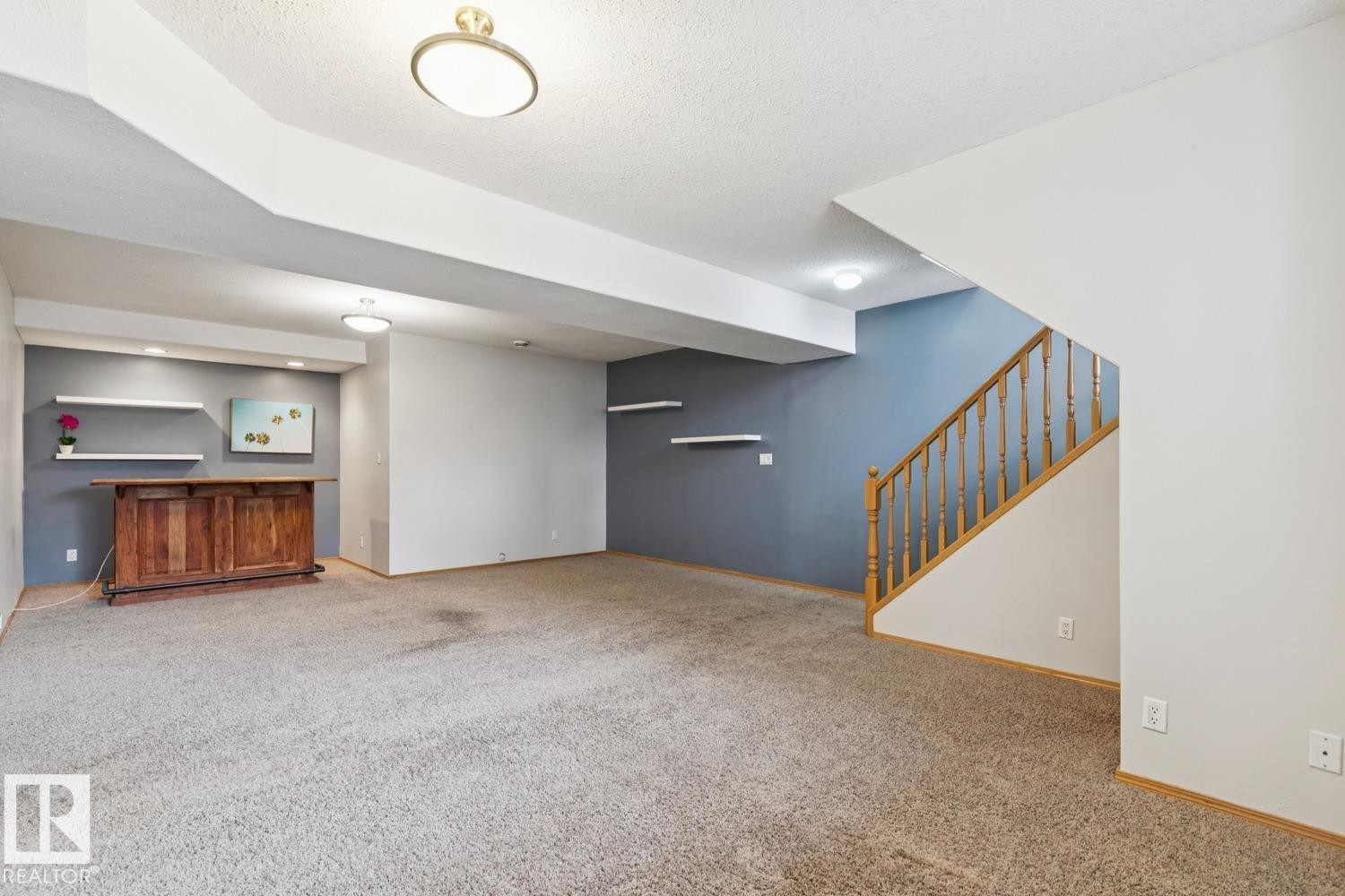 Spacious carpeted room featuring a built-in wooden bar, recessed lighting, floating wall shelves, and a wooden staircase with balusters - 1 225 Blackburn Drive E, Edmonton, AB