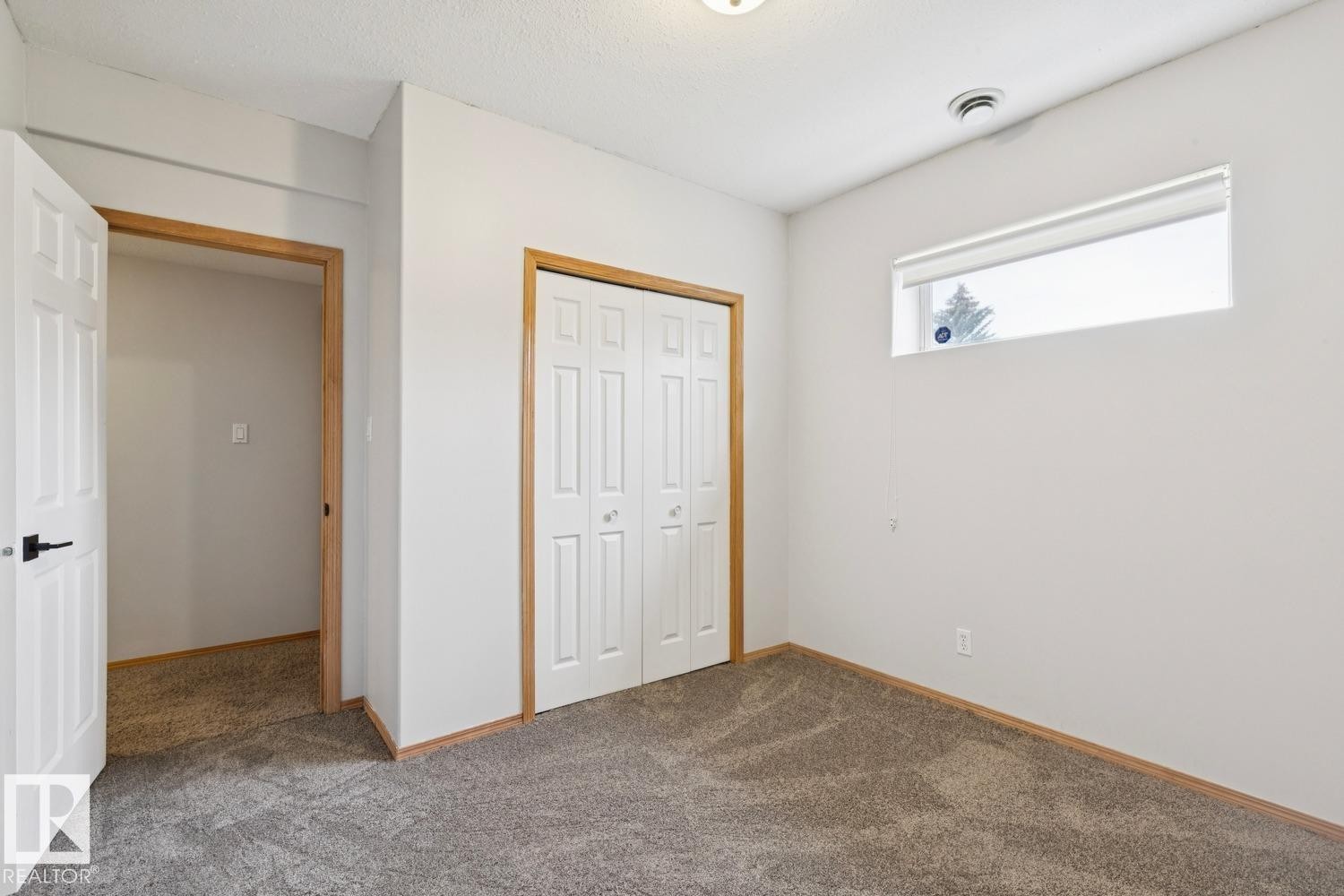 Carpeted room featuring a horizontal window with a roller shade, white bi-fold closet doors, and wood-trim doorways - 1 225 Blackburn Drive E, Edmonton, AB