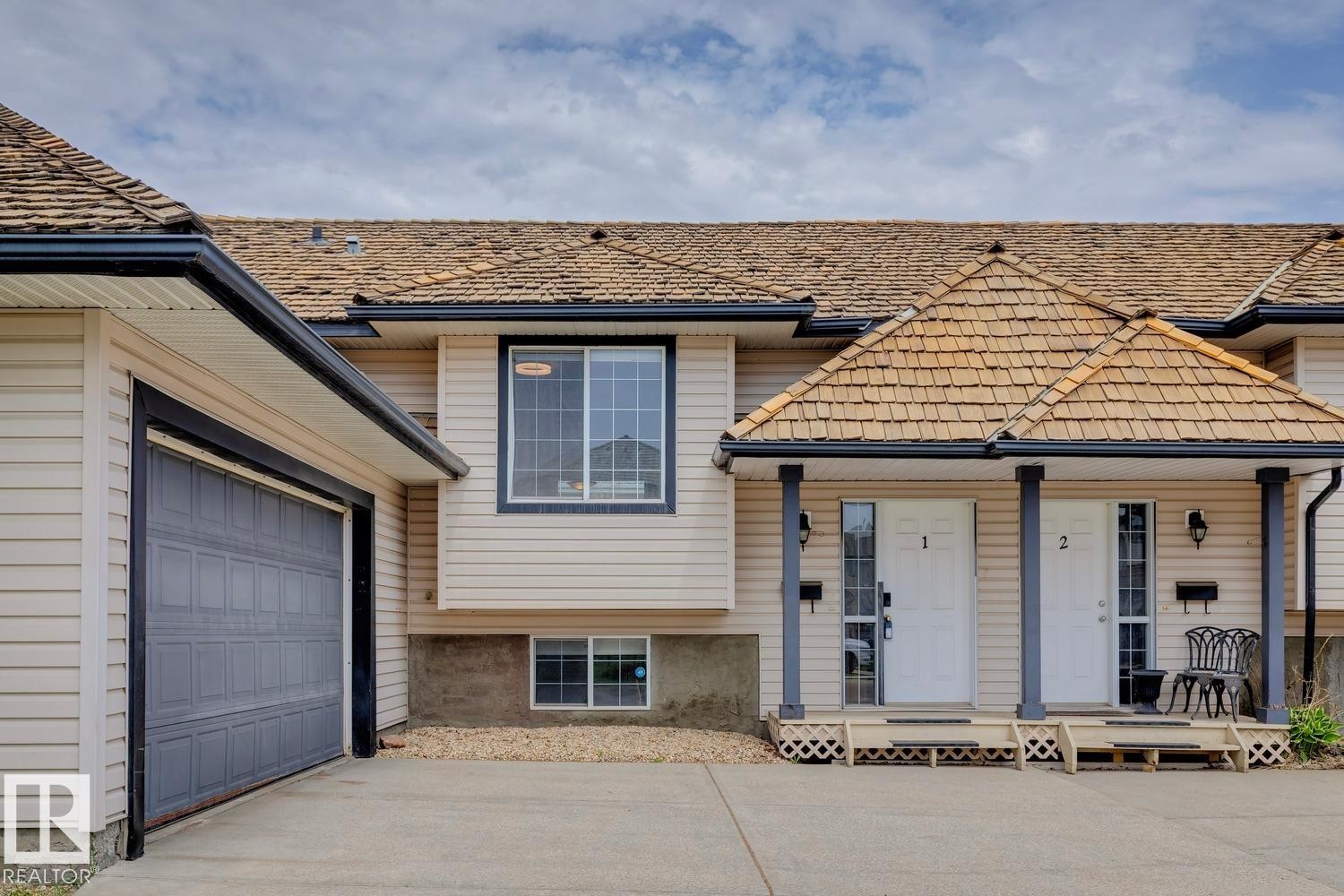 Exterior featuring light-colored siding, a dark gray paneled garage door, and a cedar shake roof - 1 225 Blackburn Drive E, Edmonton, AB