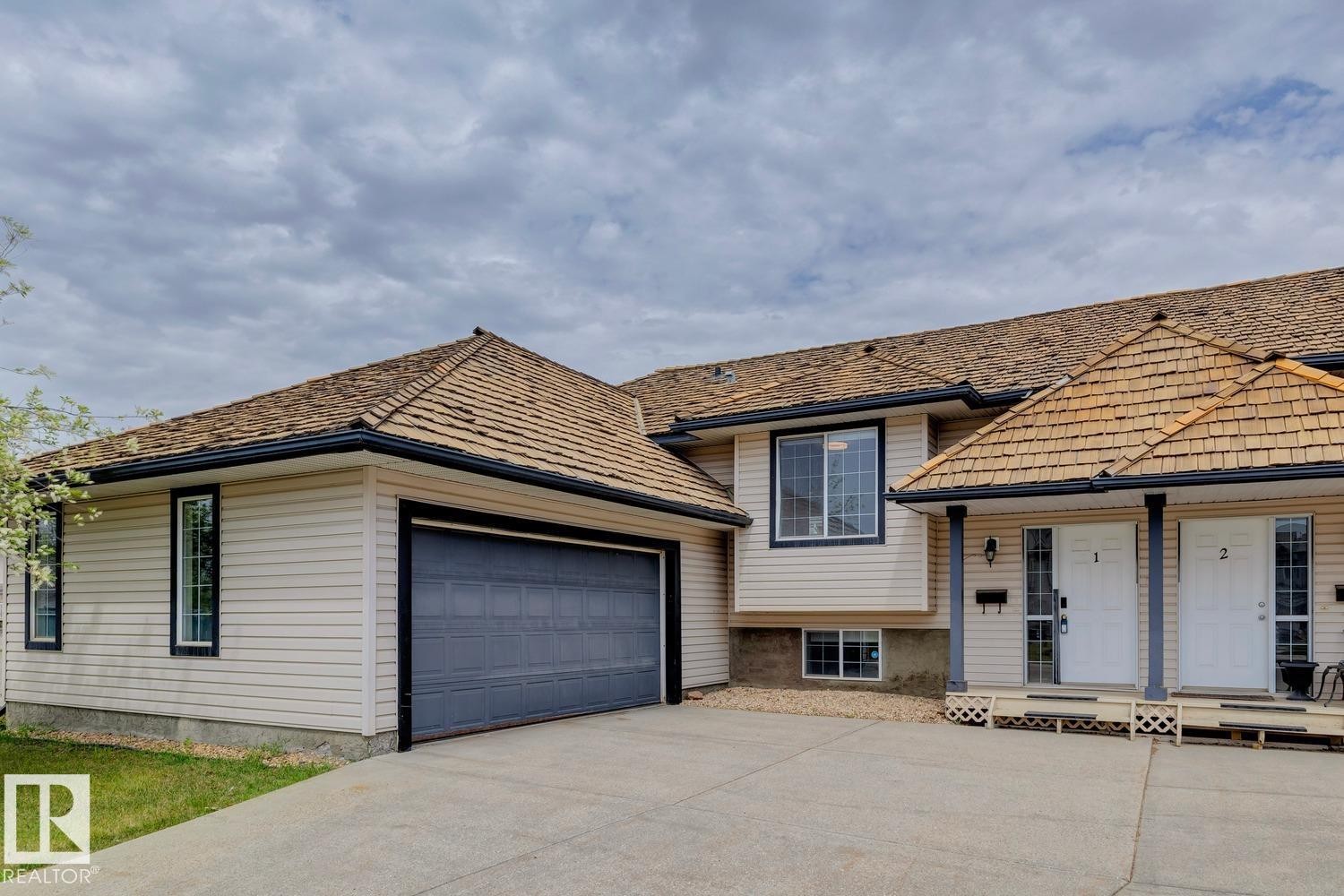 Duplex exterior featuring light beige siding, a two-car garage with a gray door, and a cedar shake roof - 1 225 Blackburn Drive E, Edmonton, AB