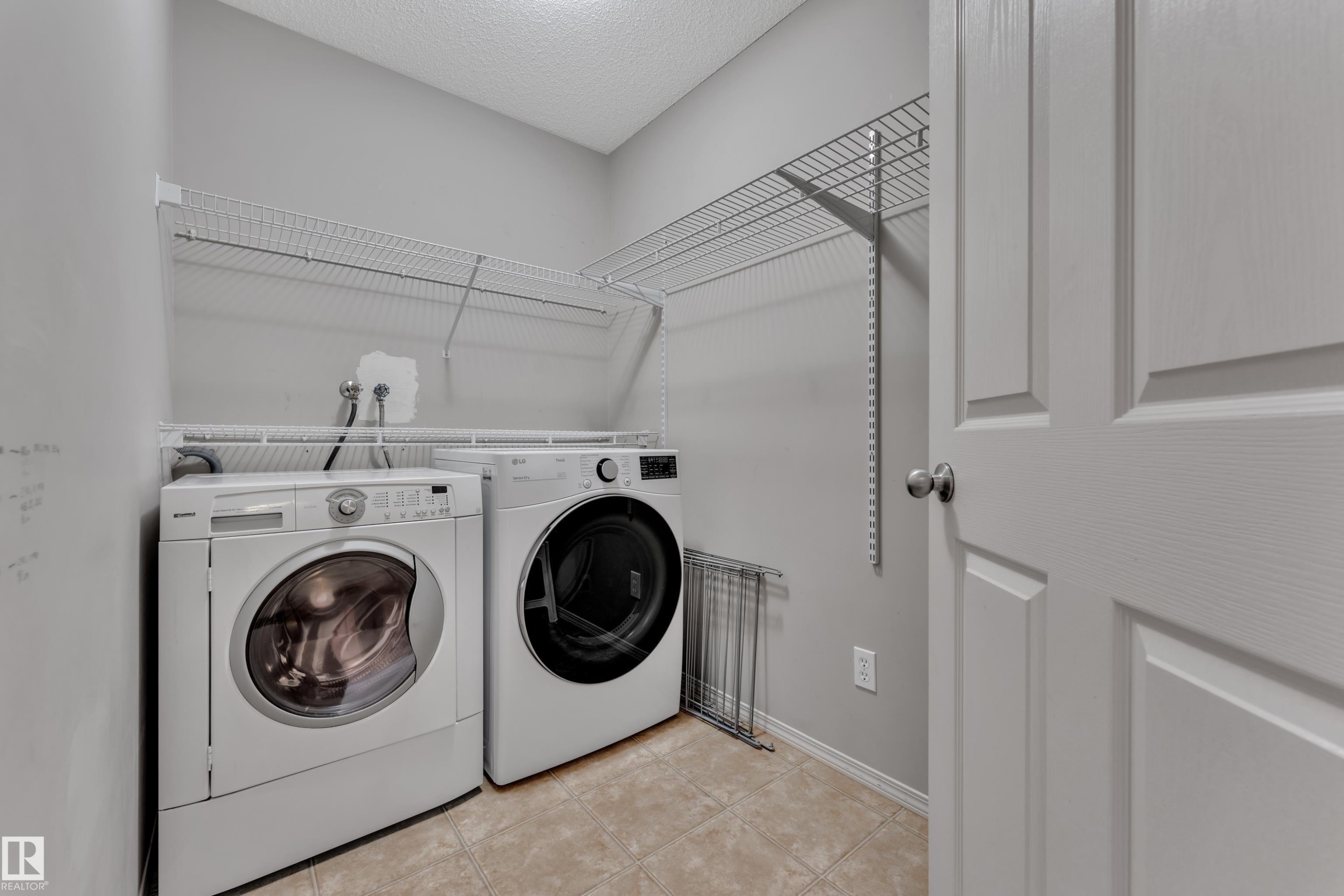 Dedicated laundry area featuring front-loading appliances, overhead wire shelving, light grey painted walls, and tan tile flooring - 14031 136 Street, Edmonton, AB