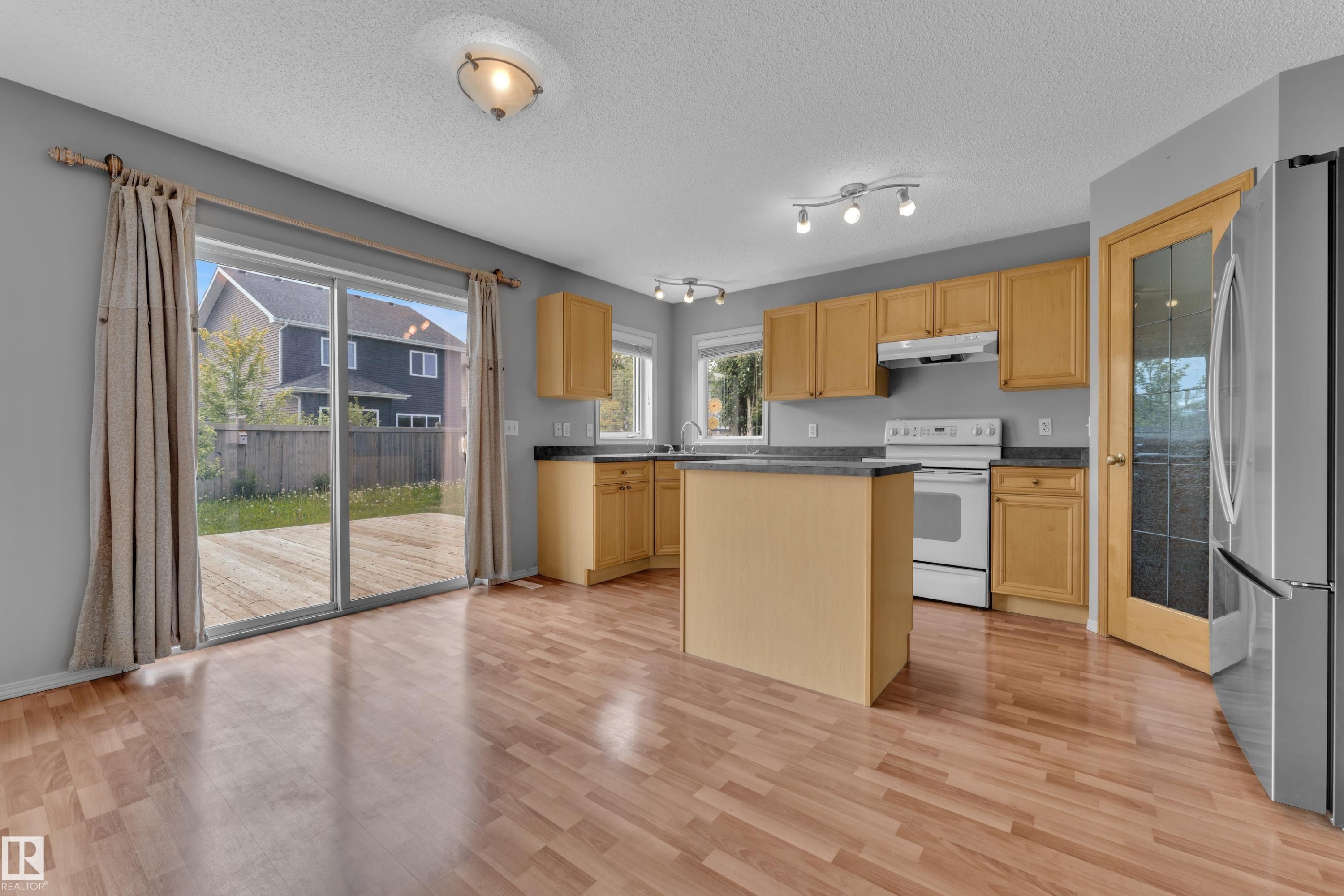 Open-concept kitchen and living area featuring wood-finish flooring, ample natural light, light wood cabinetry, a central island, and direct outdoor access via sliding glass doors to a backyard deck - 14031 136 Street, Edmonton, AB