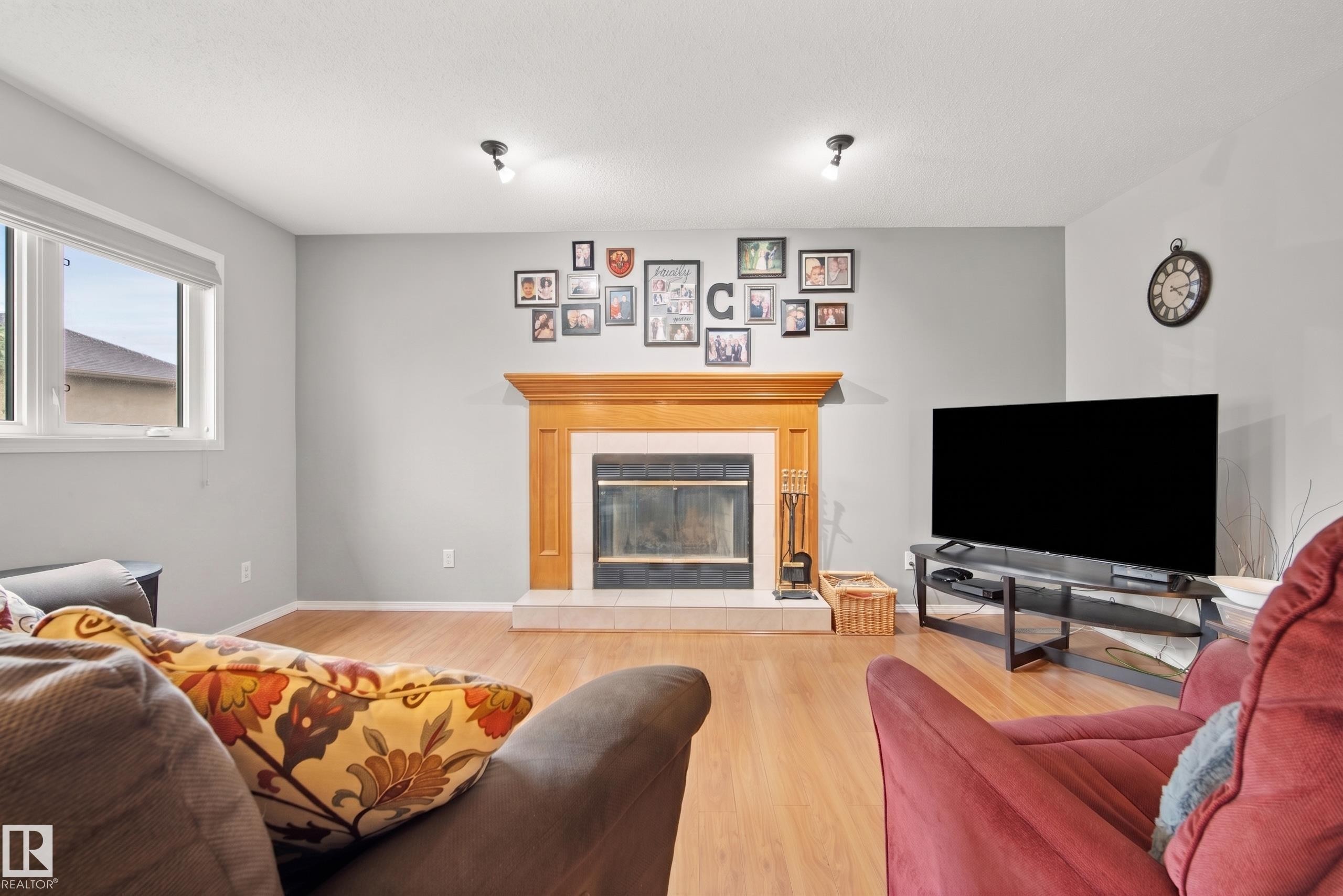 Living area with wood-finish flooring, a fireplace featuring a light wood mantel and white tile hearth, and a window with white trim - 15503 62 Street, Edmonton, AB