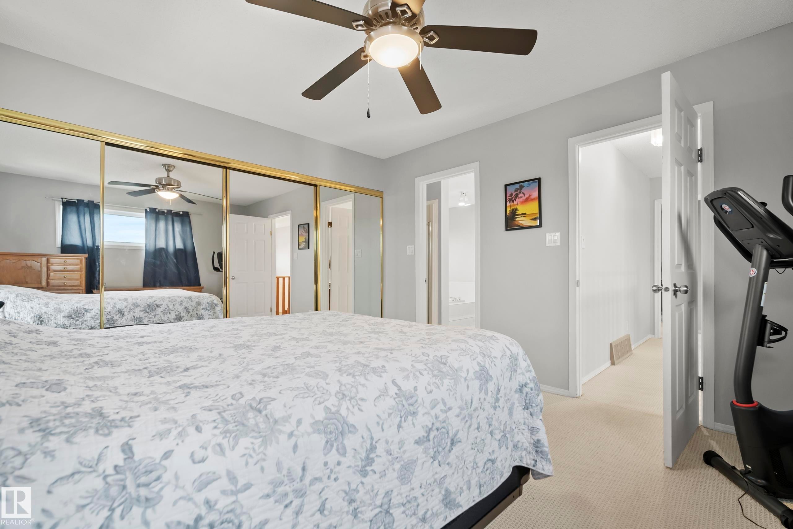 Bedroom featuring a ceiling fan with integrated lighting, mirrored sliding closet doors with gold-tone framing, light-toned carpeting, and a grey wall finish - 15503 62 Street, Edmonton, AB
