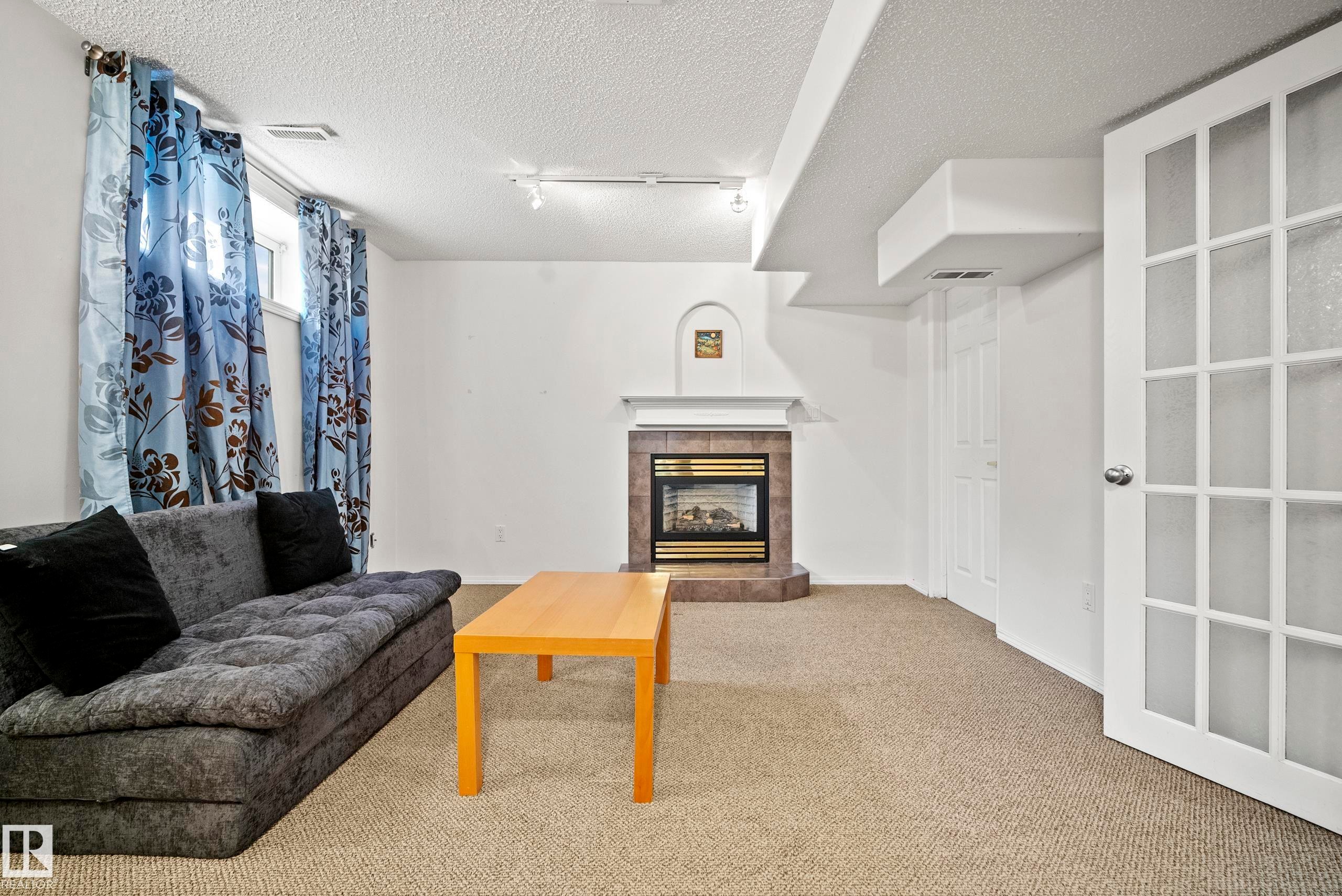 Spacious room featuring a corner fireplace with a tiled surround and white mantel, light-colored carpeting, a window with natural light, and a white French door with frosted glass panels - 15503 62 Street, Edmonton, AB