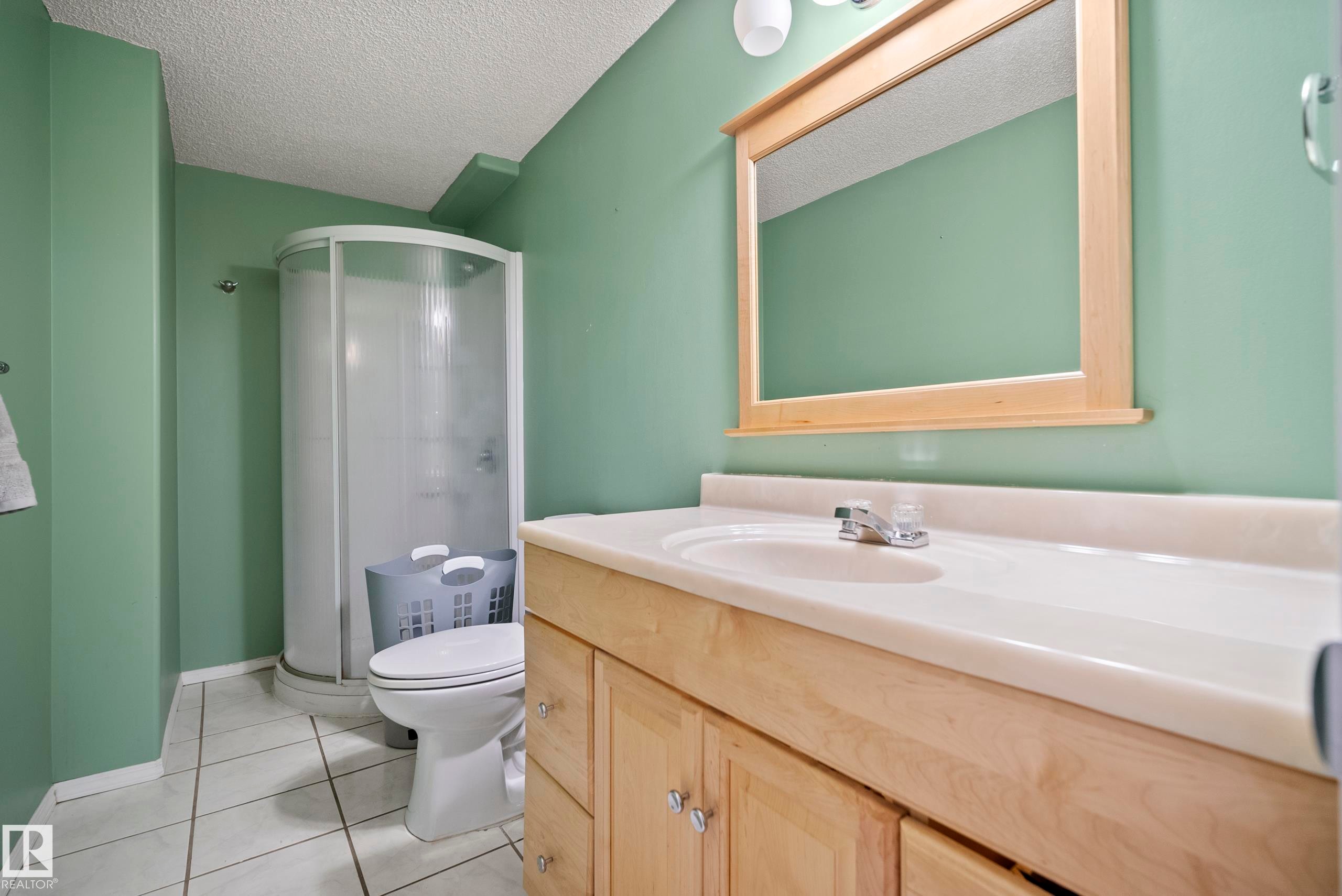 Bathroom featuring a wood-finish vanity with a built-in sink, a framed mirror, a curved shower enclosure, and light-colored tile flooring - 15503 62 Street, Edmonton, AB
