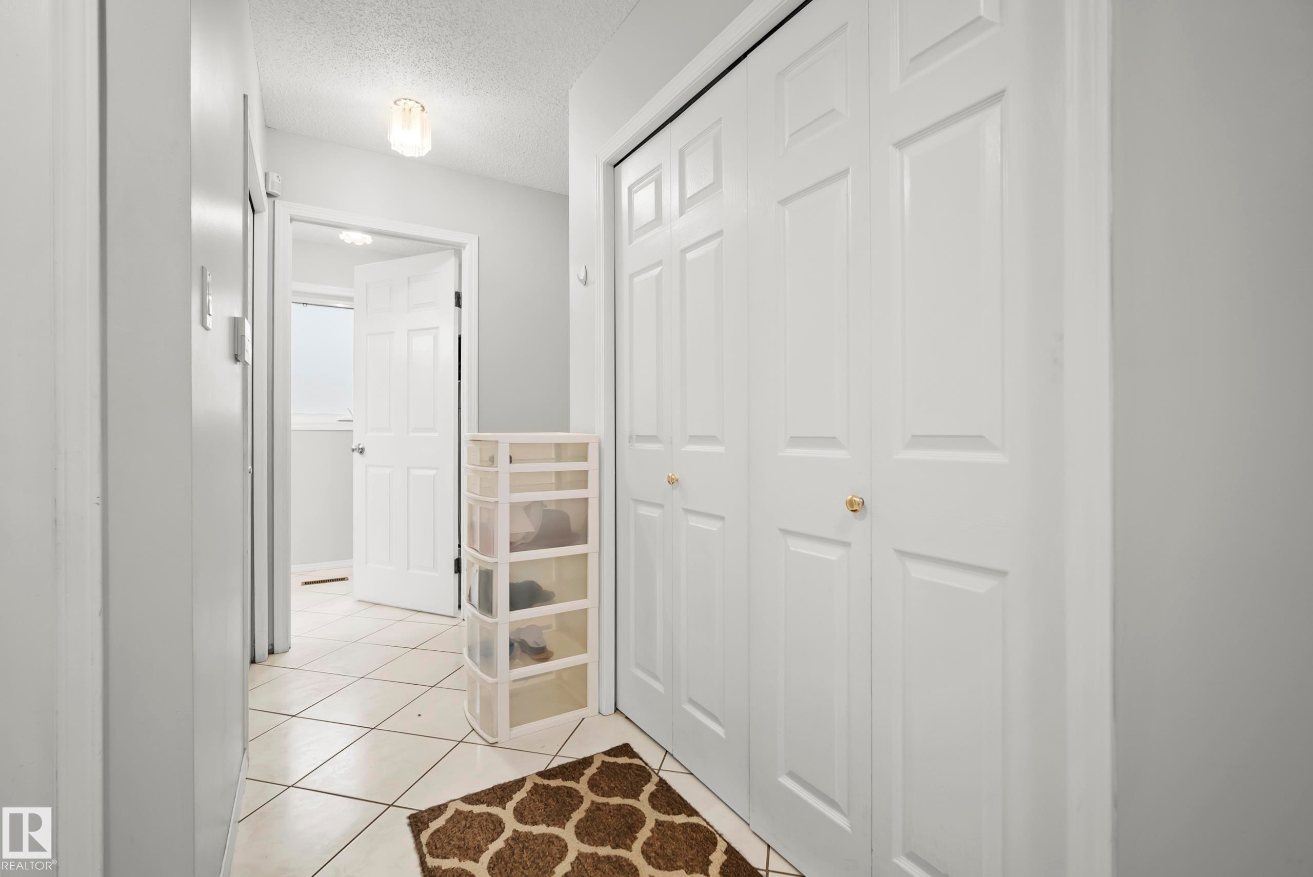 Hallway featuring light tile flooring with dark grout, white paneled bifold closet doors with gold-tone hardware, and painted walls - 15503 62 Street, Edmonton, AB