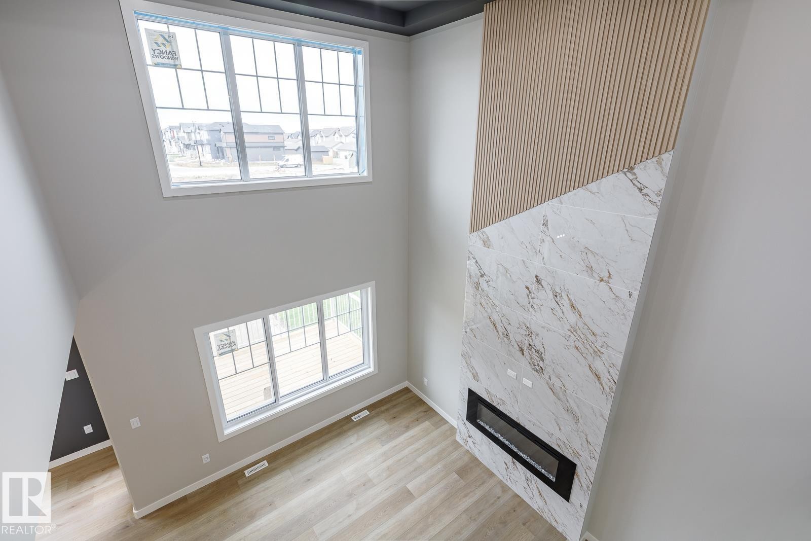 Two-story living area featuring a full-height fireplace with a linear insert, wood-finish flooring, two large windows, and a striking accent wall with slat paneling and stone-look tiling - 50 Chartres Close, St. Albert, AB