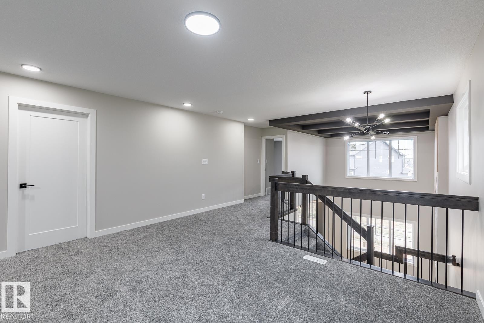 Carpeted upper-level hall featuring a dark wood-finish coffered ceiling, a contemporary chandelier, and a black metal spindle railing - 50 Chartres Close, St. Albert, AB