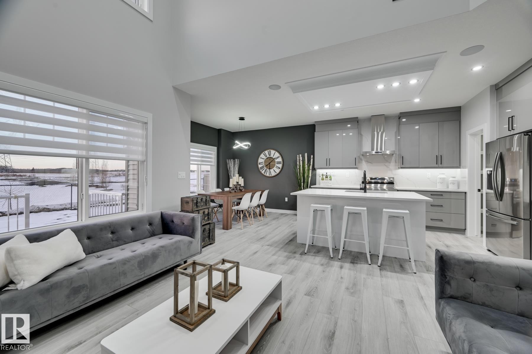 Open-concept living space featuring wood-finish flooring, a two-story ceiling, a contemporary kitchen island with a white countertop, recessed lighting, and a stainless steel refrigerator - 54 Chartres Close, St. Albert, AB - Indoor