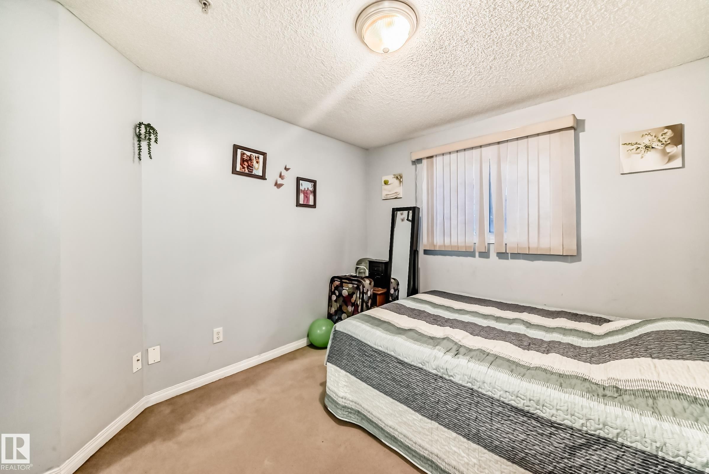 Carpeted bedroom with a single window featuring vertical blinds, light gray walls, white baseboards, and a semi-flush mount ceiling light - 108 10535 122 Street, Edmonton, AB