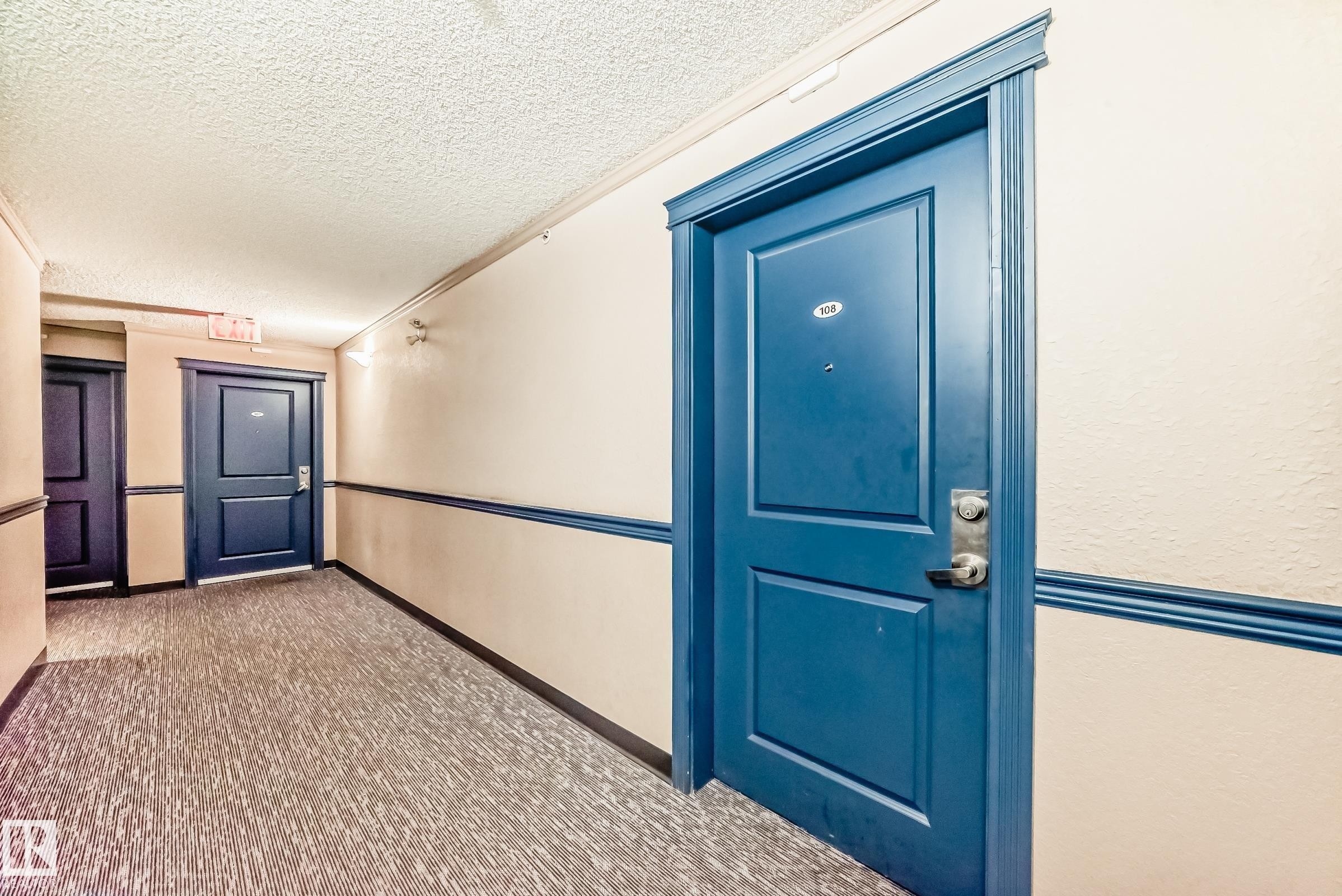 Corridor featuring textured carpeting, neutral wall finishes, and contrasting blue doors with trim - 108 10535 122 Street, Edmonton, AB