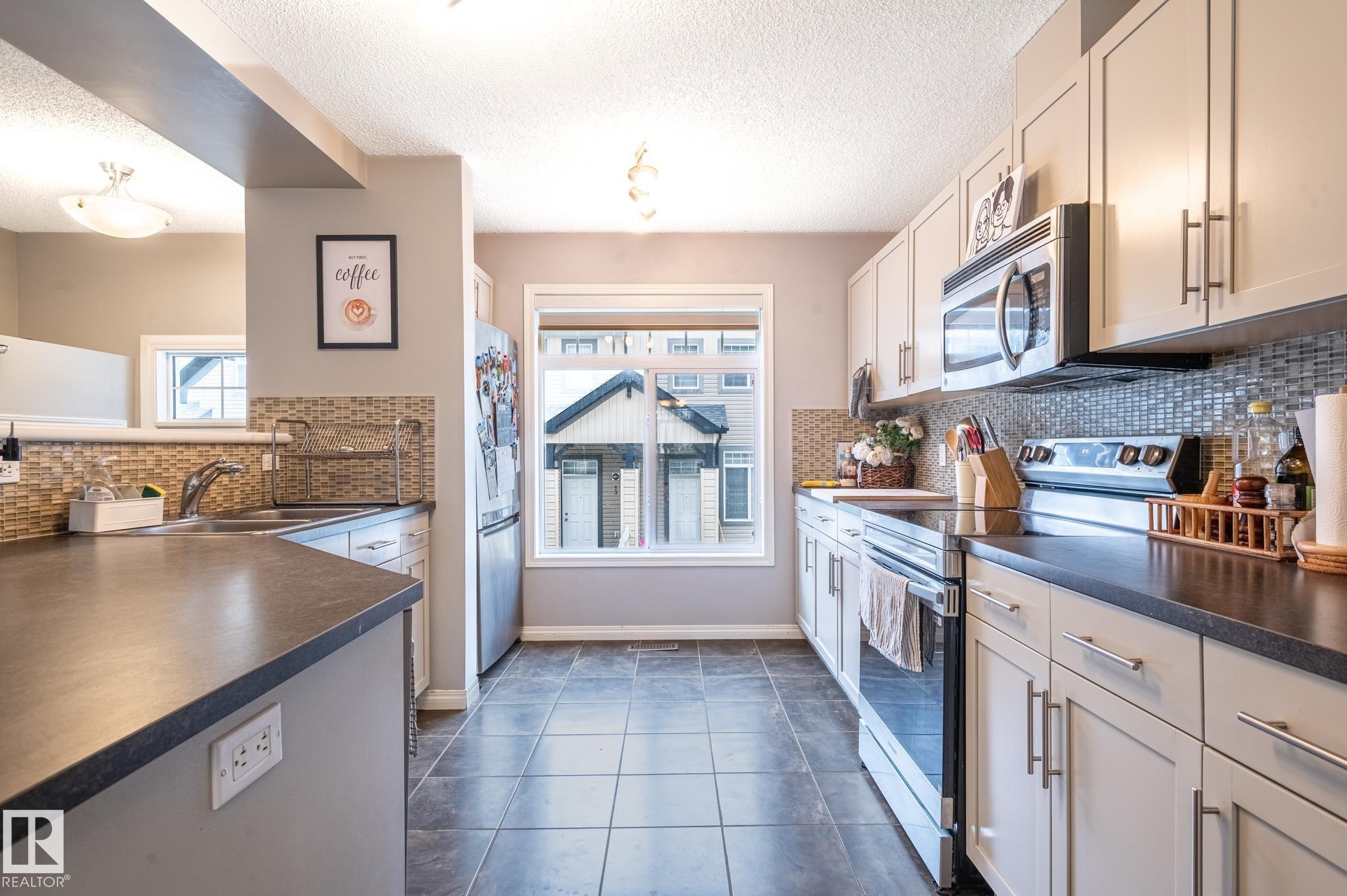 U-shaped kitchen featuring light-toned cabinetry with bar pull hardware, dark countertops, a mosaic tile backsplash, and stainless steel appliances - 46 465 Hemingway Road, Edmonton, AB