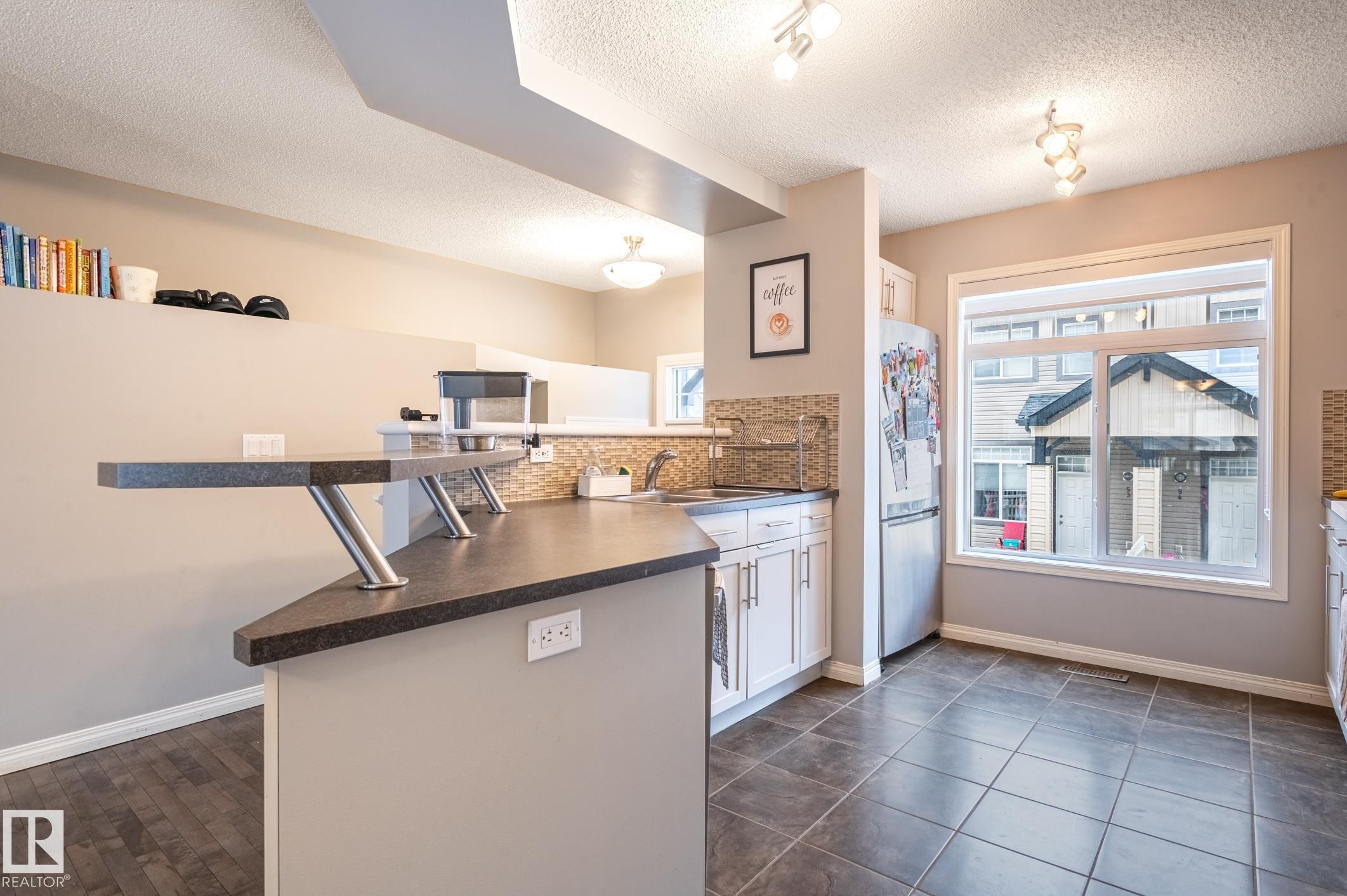Kitchen featuring a peninsula with a raised countertop and seating area, stainless steel refrigerator, white cabinetry, tile backsplash, and a large window - 46 465 Hemingway Road, Edmonton, AB