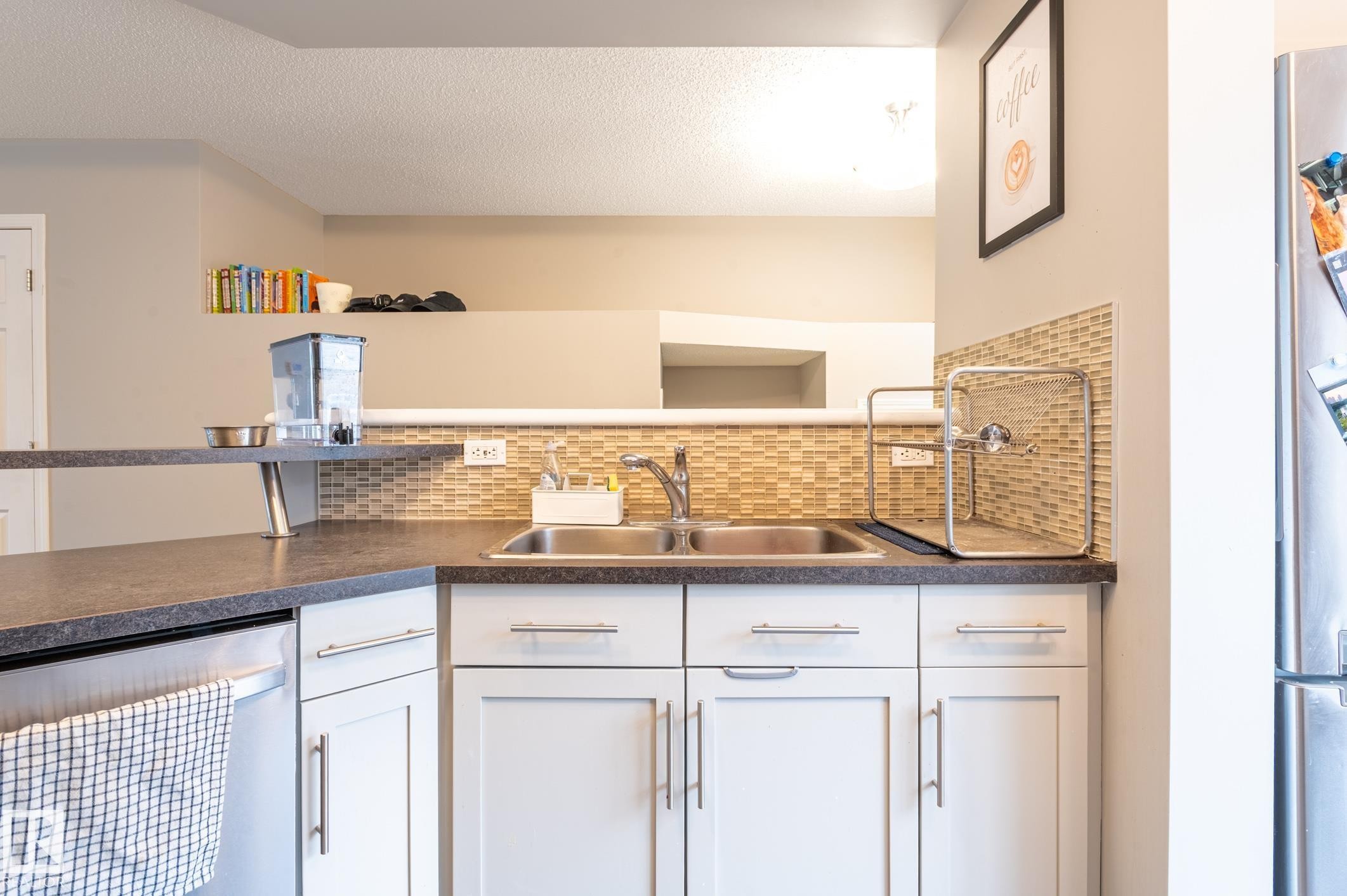 Kitchen featuring white cabinetry with brushed nickel hardware, stainless steel double basin sink, and a multi-toned mosaic tile backsplash - 46 465 Hemingway Road, Edmonton, AB