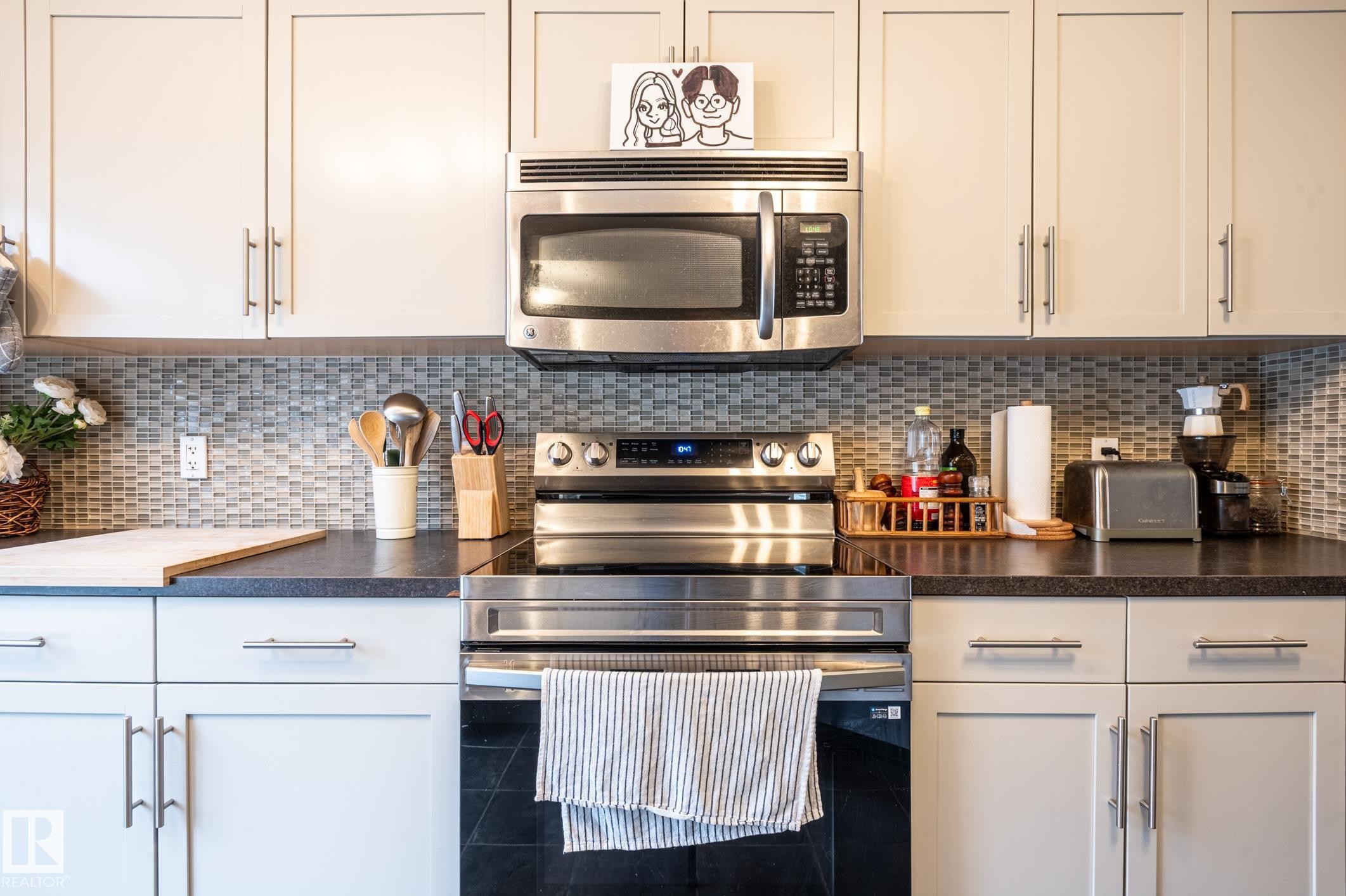 Contemporary kitchen featuring light-toned cabinetry with brushed nickel hardware, stainless steel appliances, dark countertops, and a mosaic tile backsplash - 46 465 Hemingway Road, Edmonton, AB