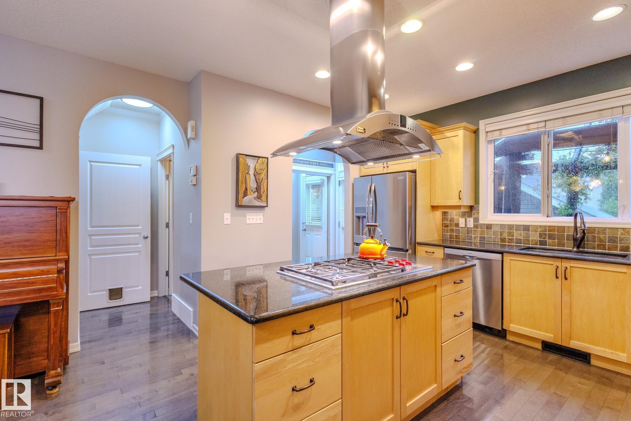 Kitchen featuring a central island with gas cooktop, stainless steel range hood, stainless steel appliances, wood-finish flooring, and light-toned cabinetry with dark countertops - 7207 14A Avenue, Edmonton, AB - Indoor Photo Showing Kitchen