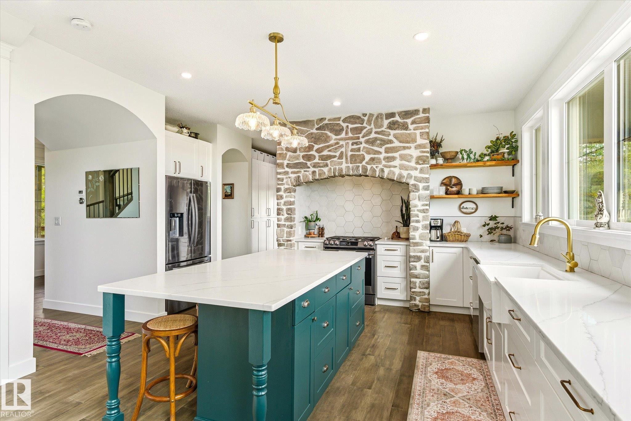 Spacious kitchen featuring a central island with a white countertop and teal cabinetry, a stone-clad range hood, white shaker-style perimeter cabinets, and a white farmhouse sink with a gold-finish faucet - 59102 Rge Road 234, Rural Westlock County, AB