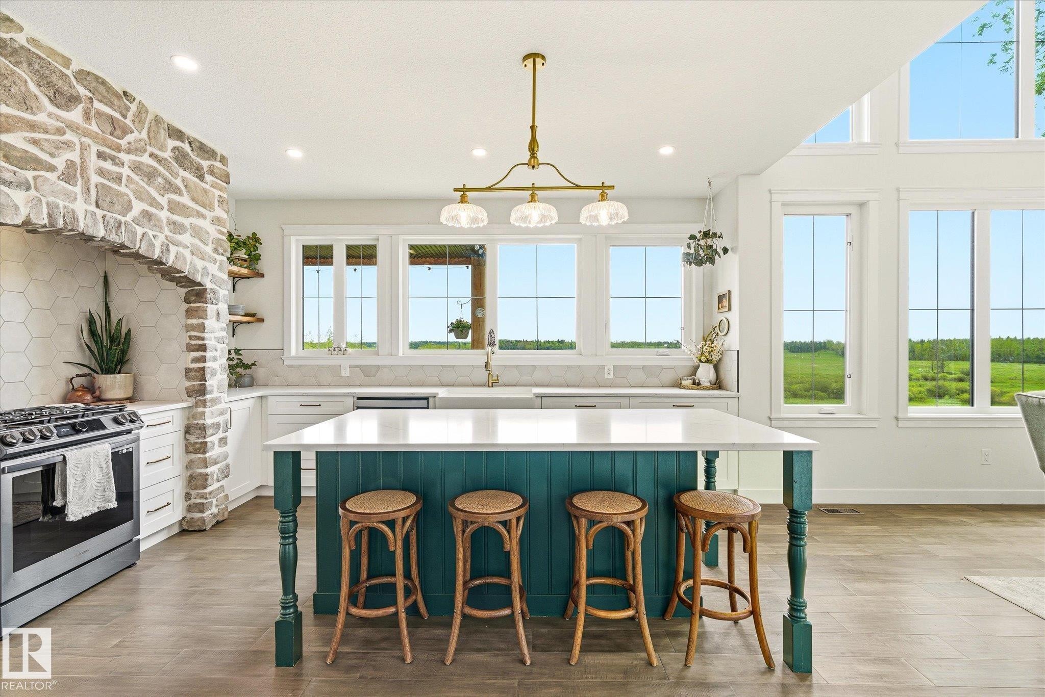 Open-concept kitchen featuring a teal island with a white countertop, stainless steel range, custom stone-finish range hood, wood-finish flooring, and a multi-window wall - 59102 Rge Road 234, Rural Westlock County, AB
