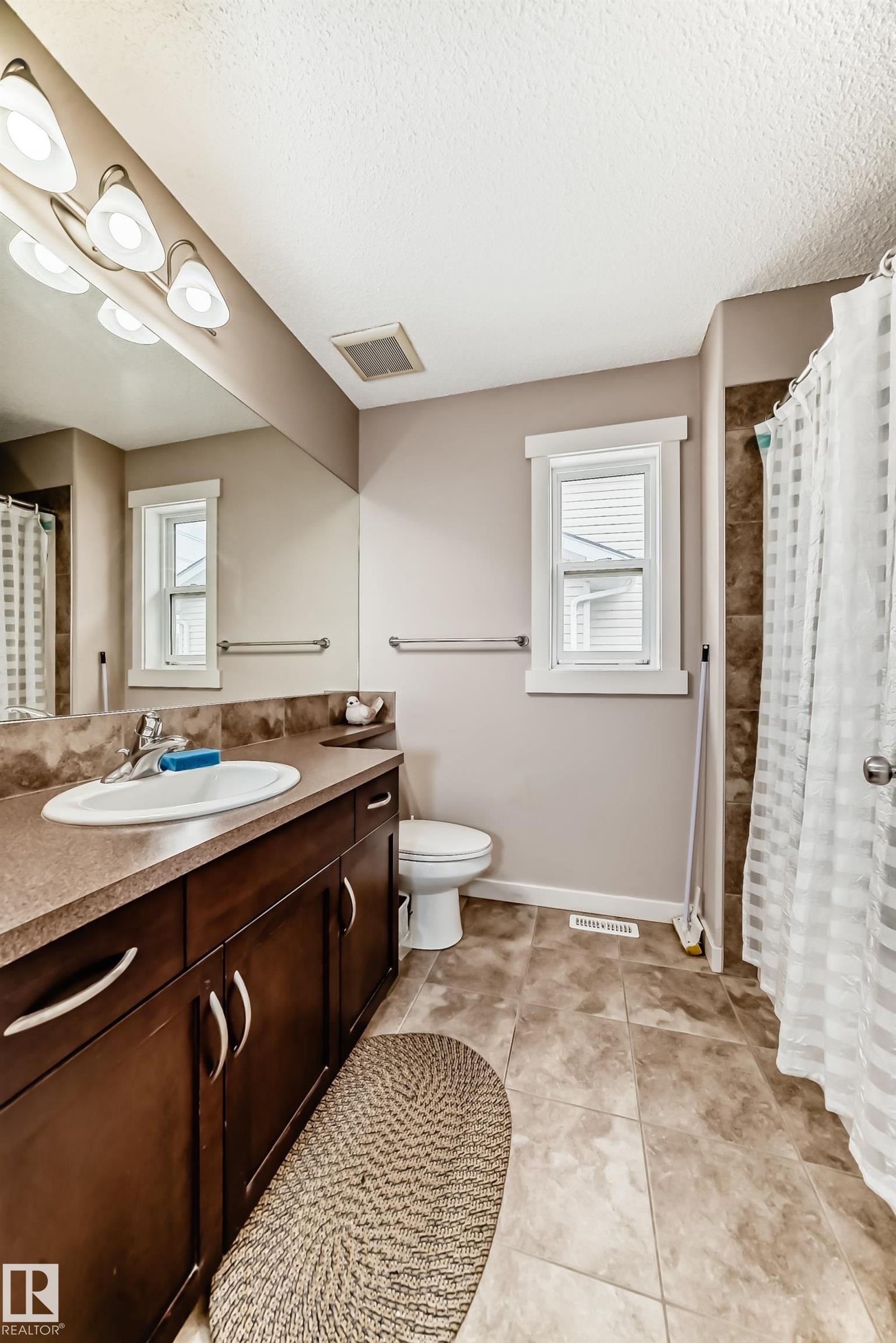 Full bathroom featuring a dark wood-finish vanity with a single basin sink, a large wall-mounted mirror, ceramic tile flooring, and a window with white trim - 2808 36 Avenue, Edmonton, AB