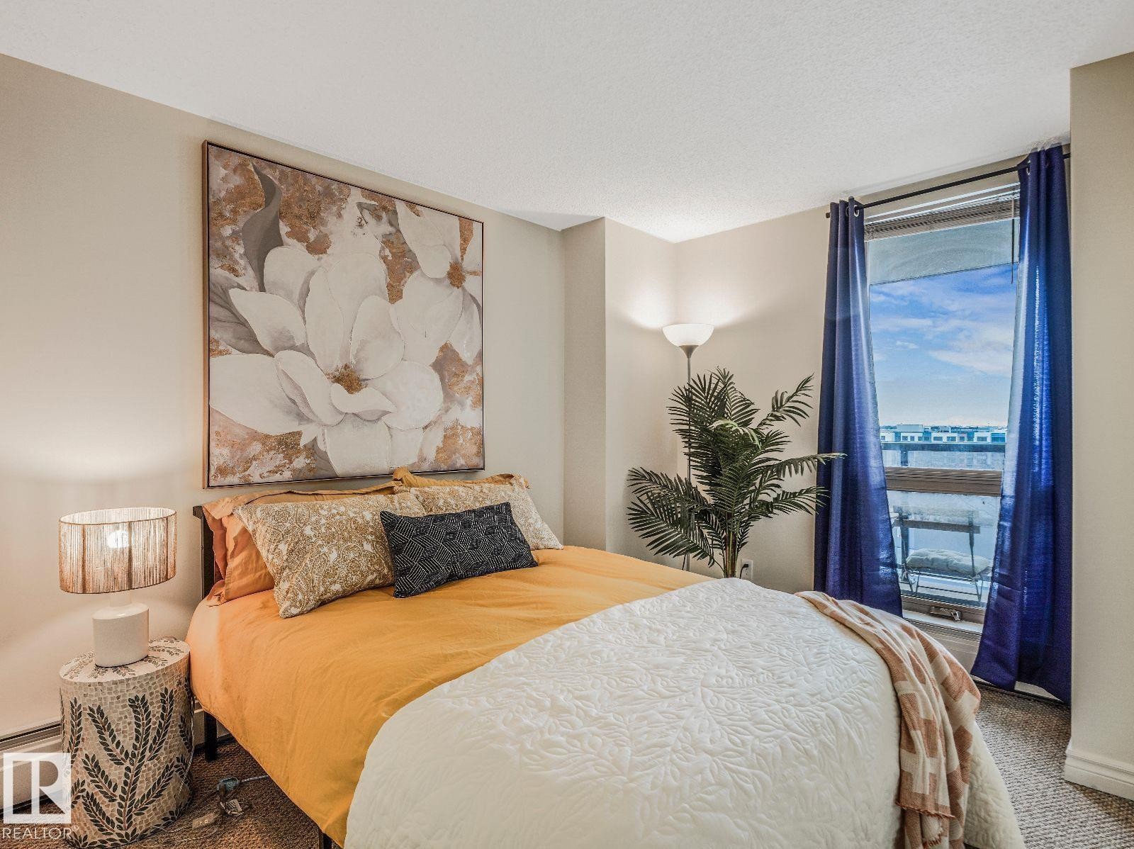 Carpeted bedroom featuring a large window with blue drapery, a floor lamp, and a modern wall art piece - 806 10149 Saskatchewan Drive, Edmonton, AB