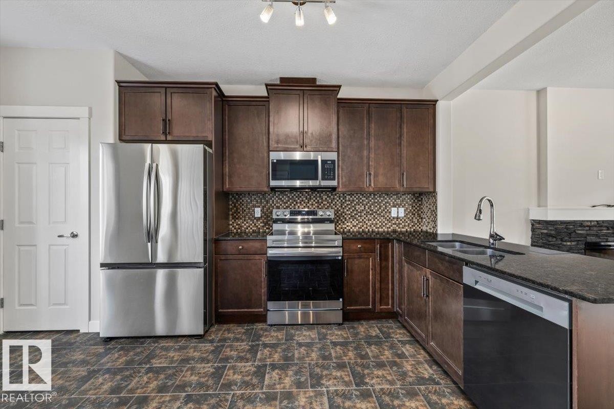 Kitchen featuring dark wood cabinetry, stainless steel appliances, a mosaic tile backsplash, dark stone countertops, and large format floor tiles - 356 Pioneer Road, Spruce Grove, AB