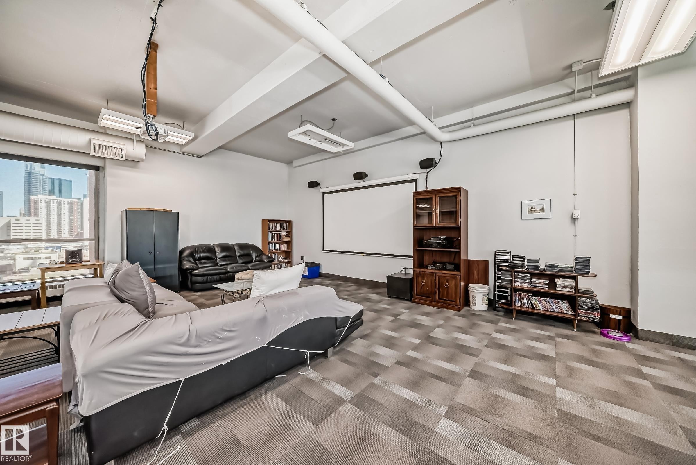 Spacious common area featuring large windows, geometric pattern carpet, exposed overhead ductwork, and a built-in projection screen - #706 10125 109 St Nw, Edmonton, AB