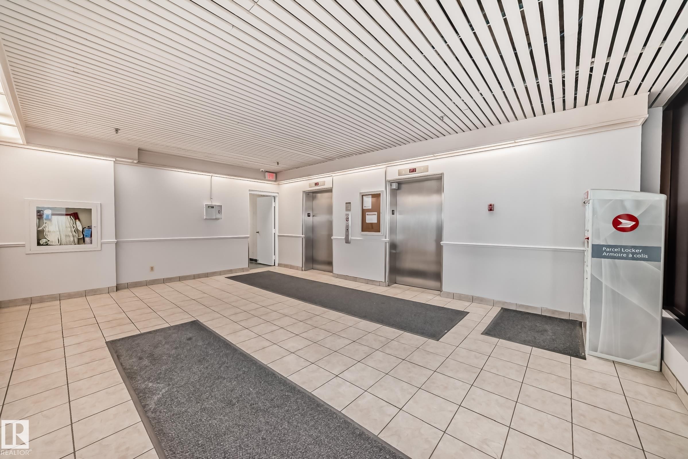 Lobby featuring two stainless steel elevators, white tile flooring, and a slatted ceiling with integrated lighting - #706 10125 109 St Nw, Edmonton, AB