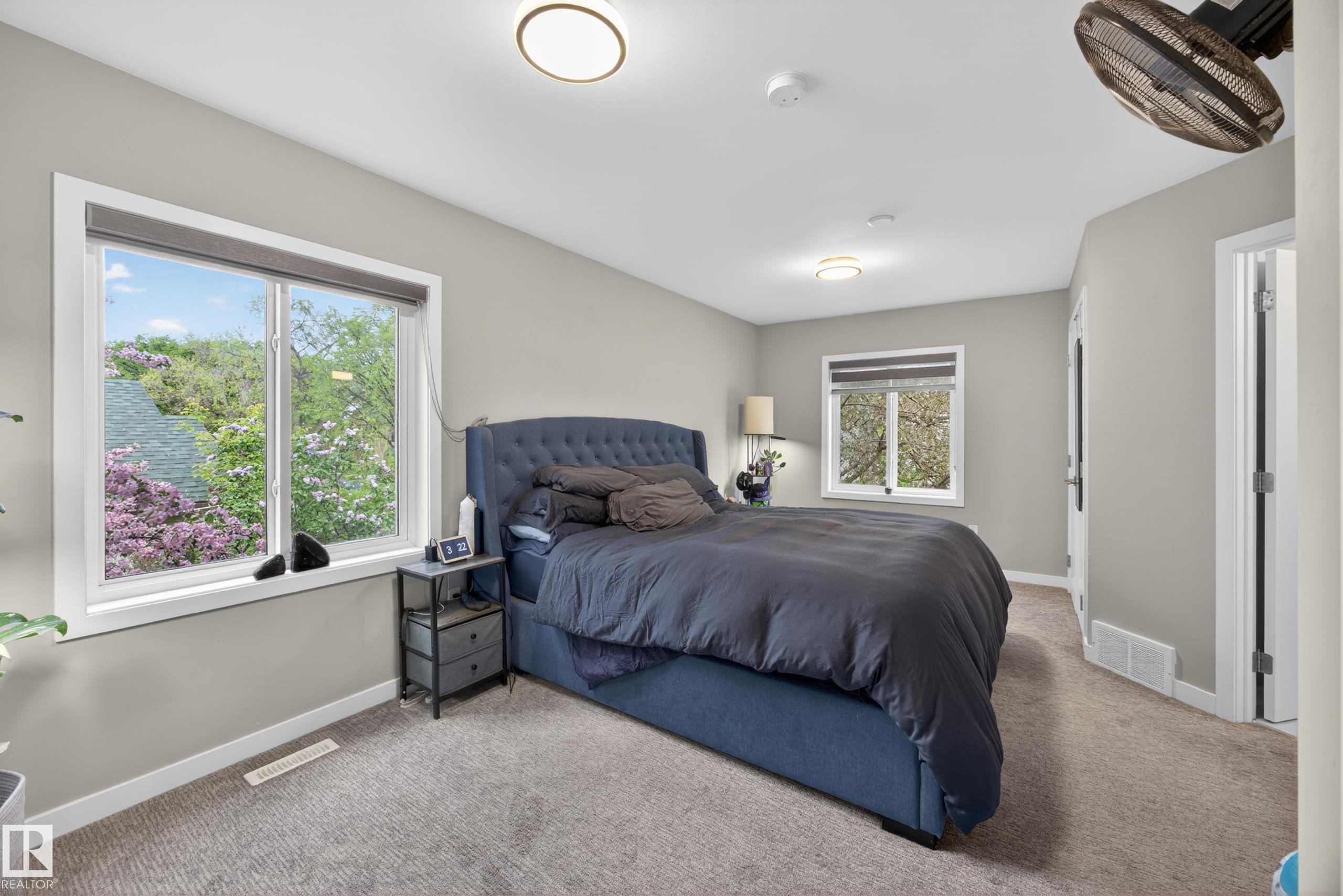Carpeted bedroom featuring two windows with roller shades, neutral painted walls, and two flush-mount ceiling lights - 12604 116 Avenue, Edmonton, AB