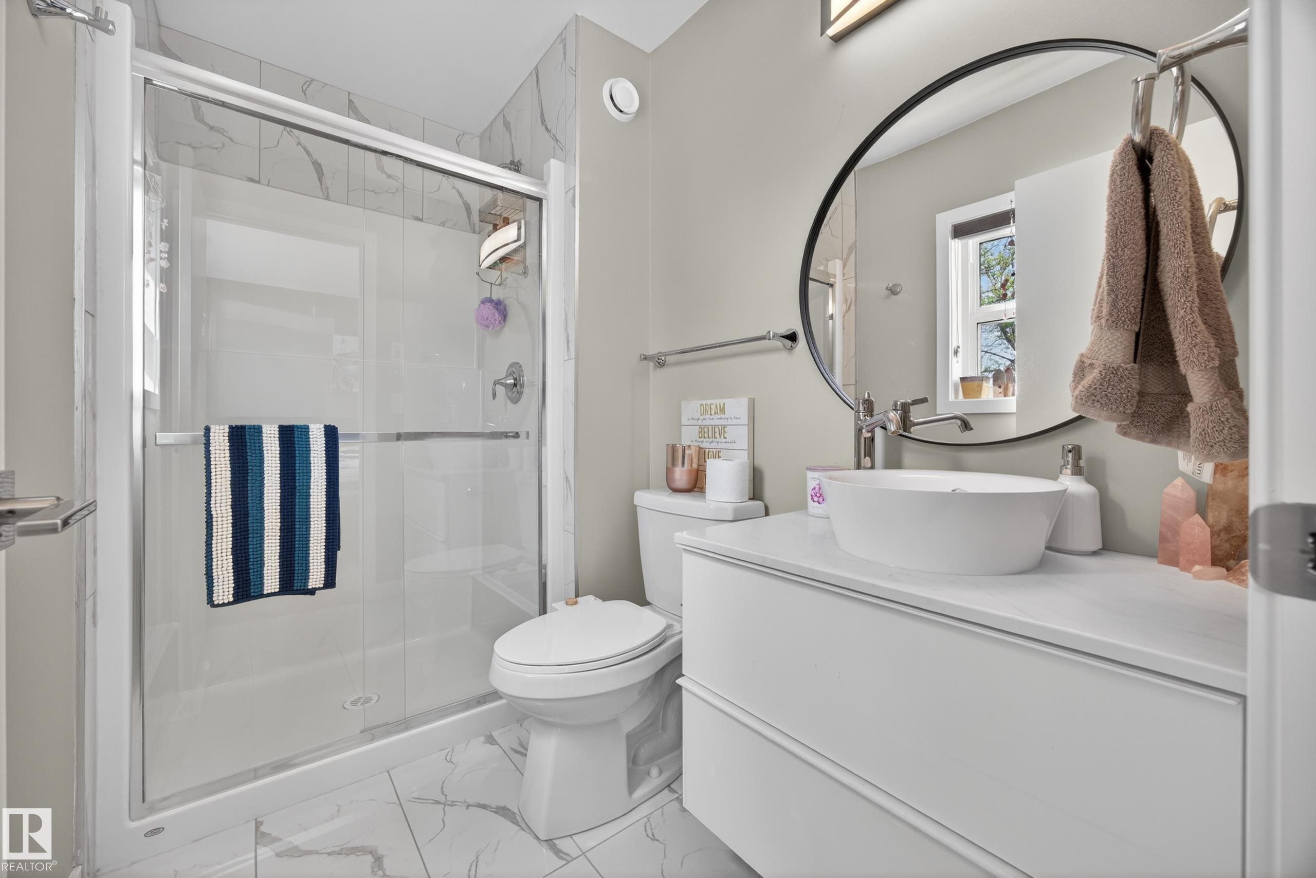Bathroom featuring a glass-enclosed shower with tile surround, a vessel sink vanity with a white countertop, and a circular wall mirror - 12604 116 Avenue, Edmonton, AB
