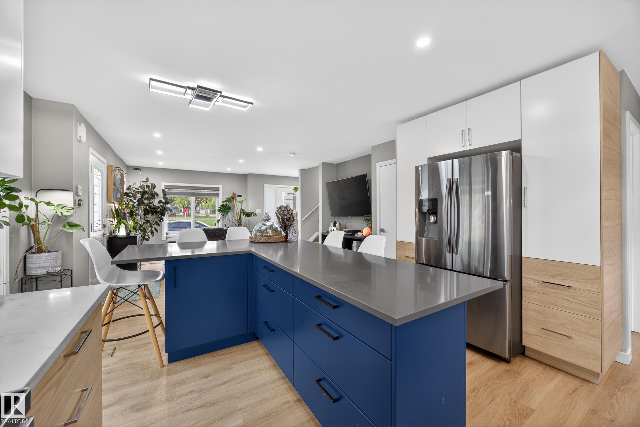 Modern kitchen featuring a blue island with a grey countertop, wood-finish flooring, recessed lighting, and white and wood-finish cabinetry surrounding a stainless steel refrigerator - 12604 116 Avenue, Edmonton, AB