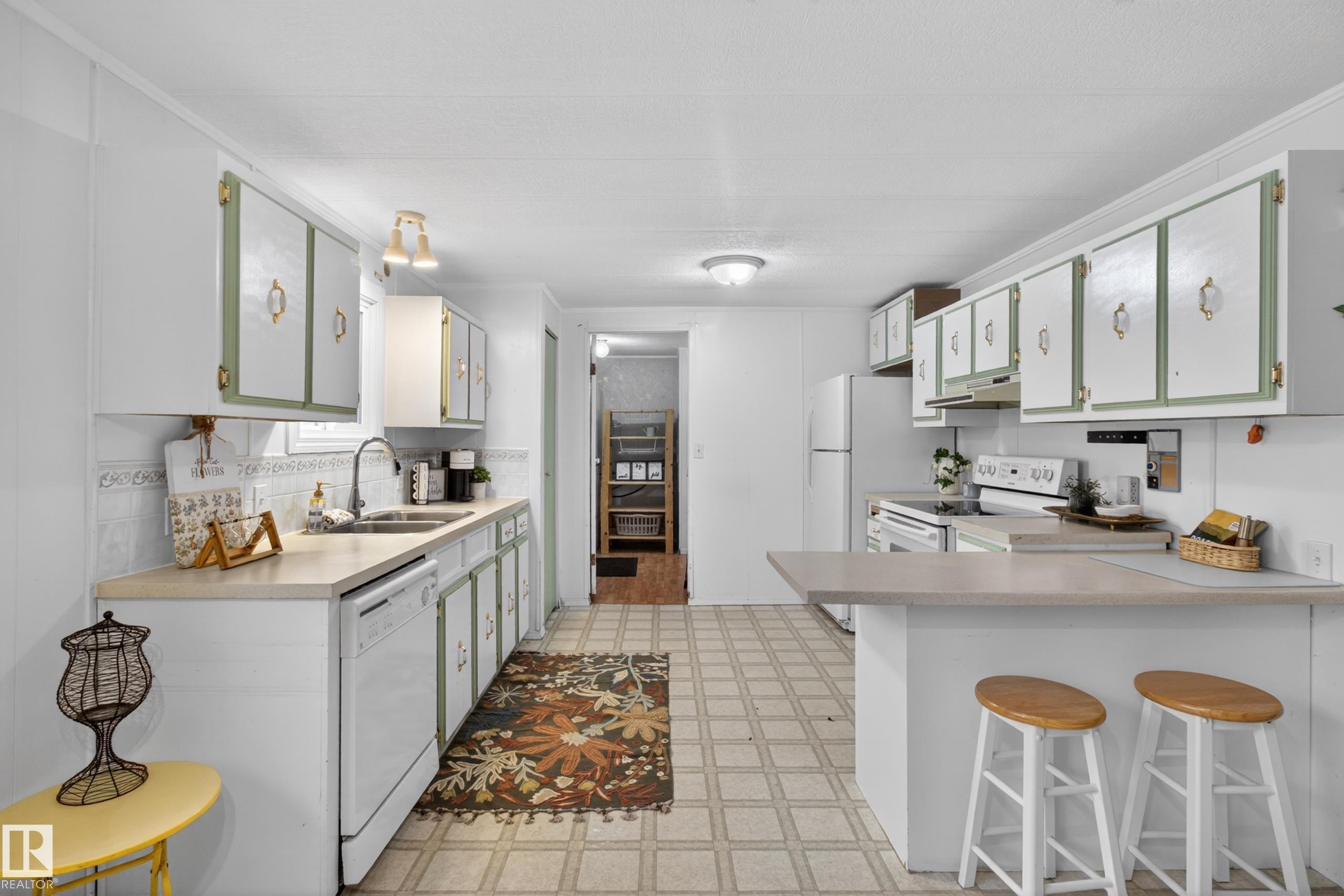 Kitchen featuring extensive white cabinetry with sage green accents and gold-tone hardware, light-colored countertops, a double basin sink with a gooseneck faucet, a white dishwasher, and a breakfast bar - 21 Willowdale Drive, Edmonton, AB - Indoor Photo Showing Kitchen With Double Sink