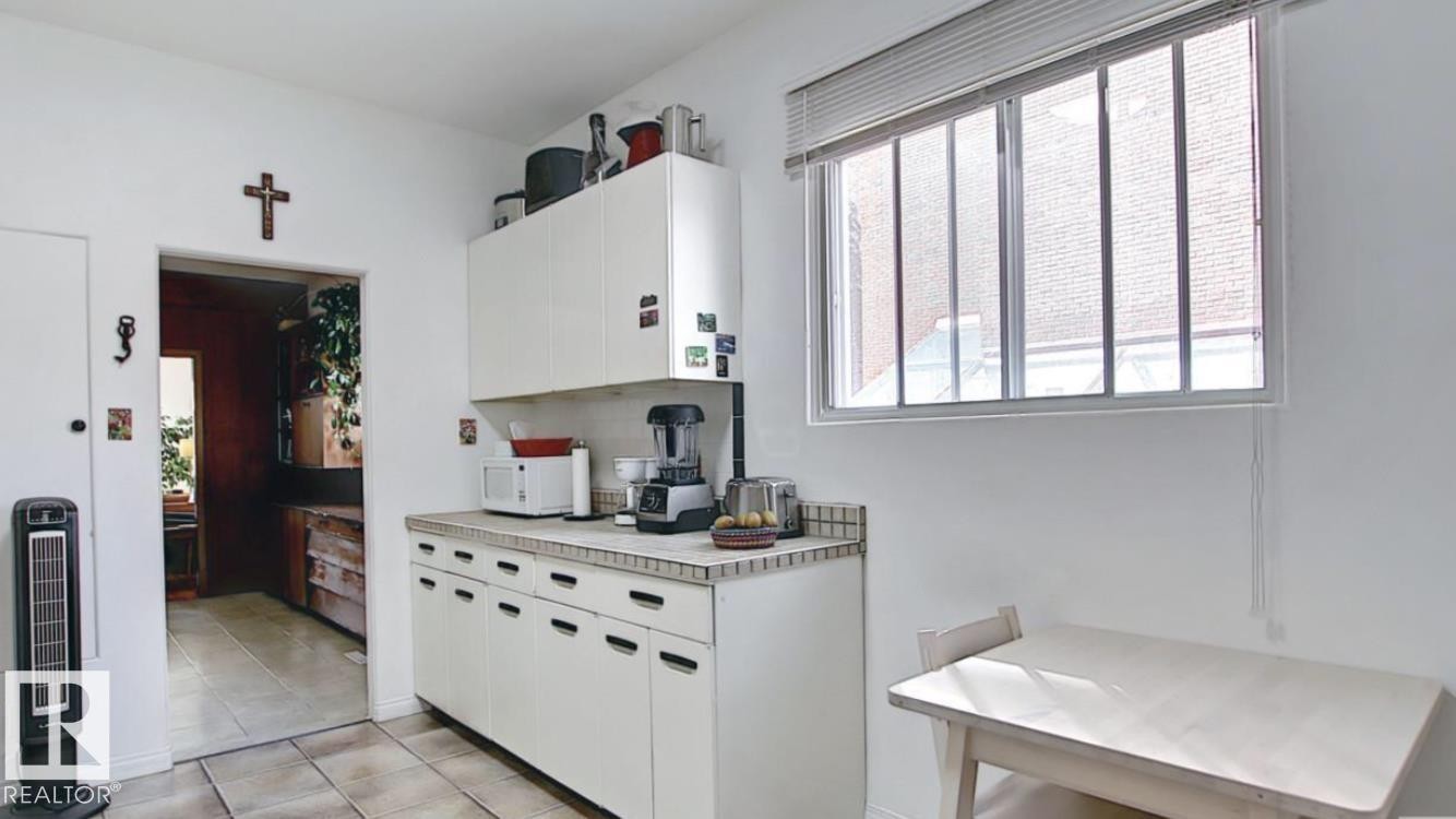 Kitchen area featuring white cabinetry with integrated pulls, a tile countertop, a window with horizontal blinds, and tile flooring - 10017 113 Street, Edmonton, AB