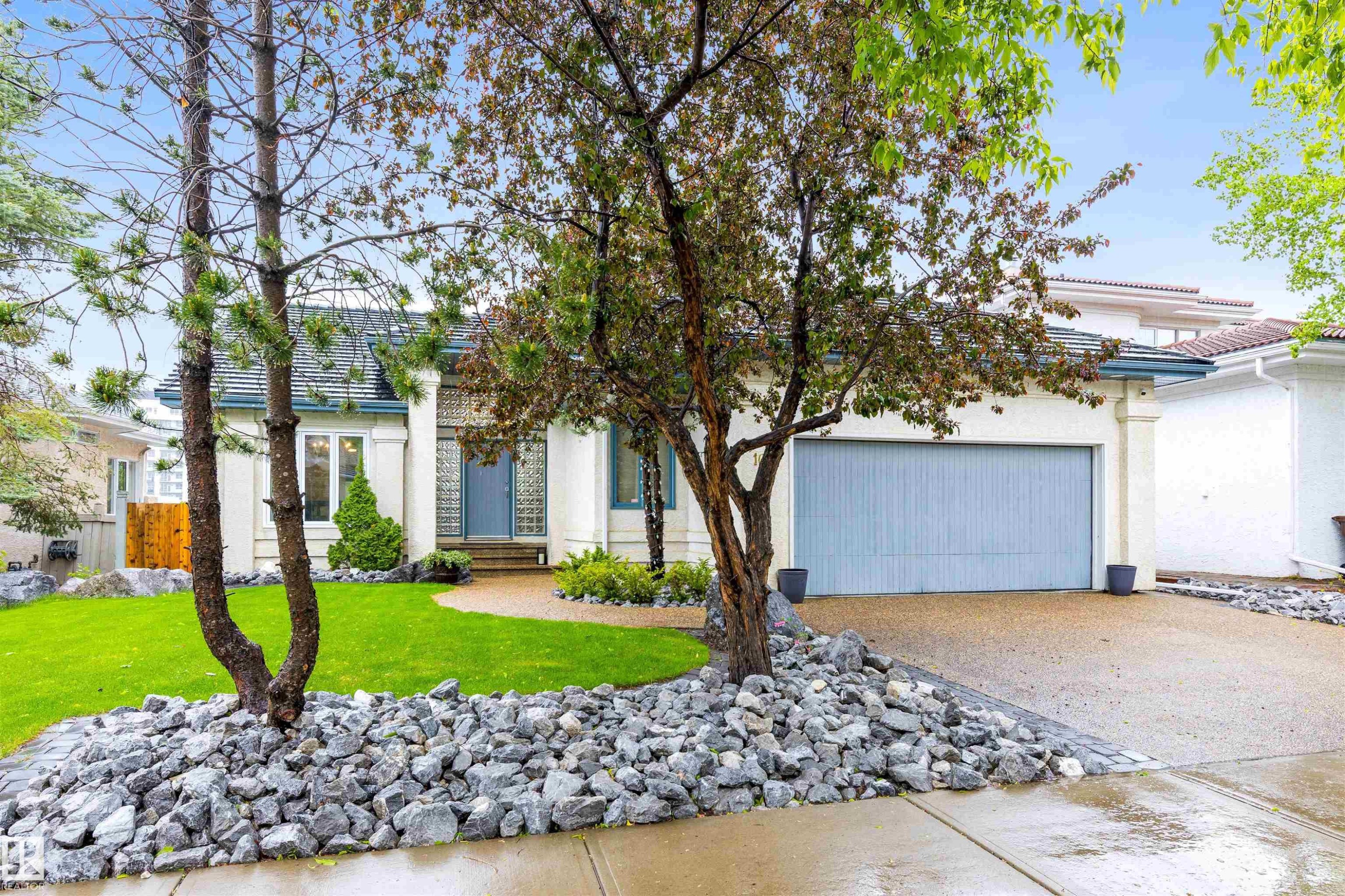 Exterior featuring a light-colored stucco finish, gray shingled roof, an attached two-car garage with a light blue door, a concrete driveway, and a front yard with manicured lawn and decorative rock landscaping - 10 Everest Crescent, St. Albert, AB