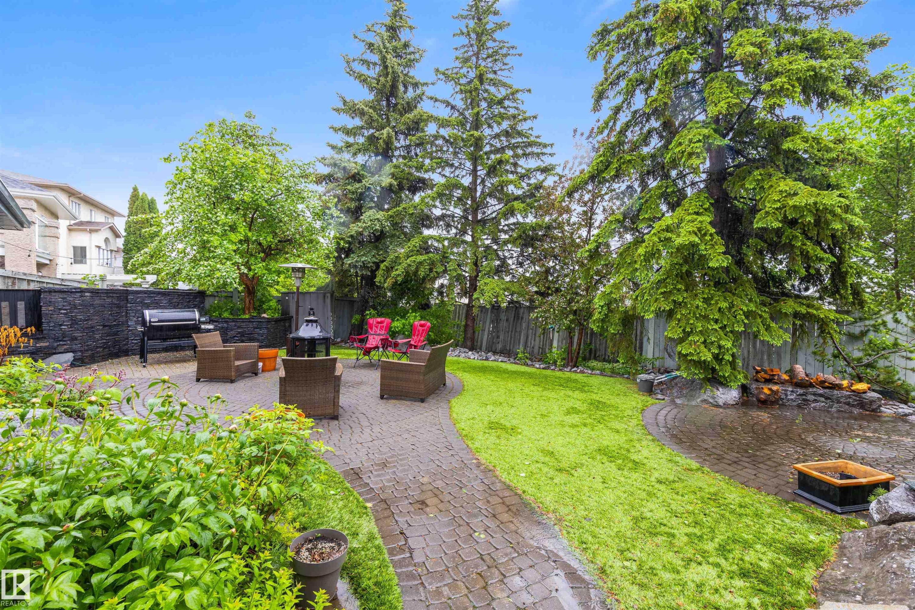 Expansive backyard featuring a paver patio, lush green lawn, mature trees, and a decorative rock feature - 10 Everest Crescent, St. Albert, AB
