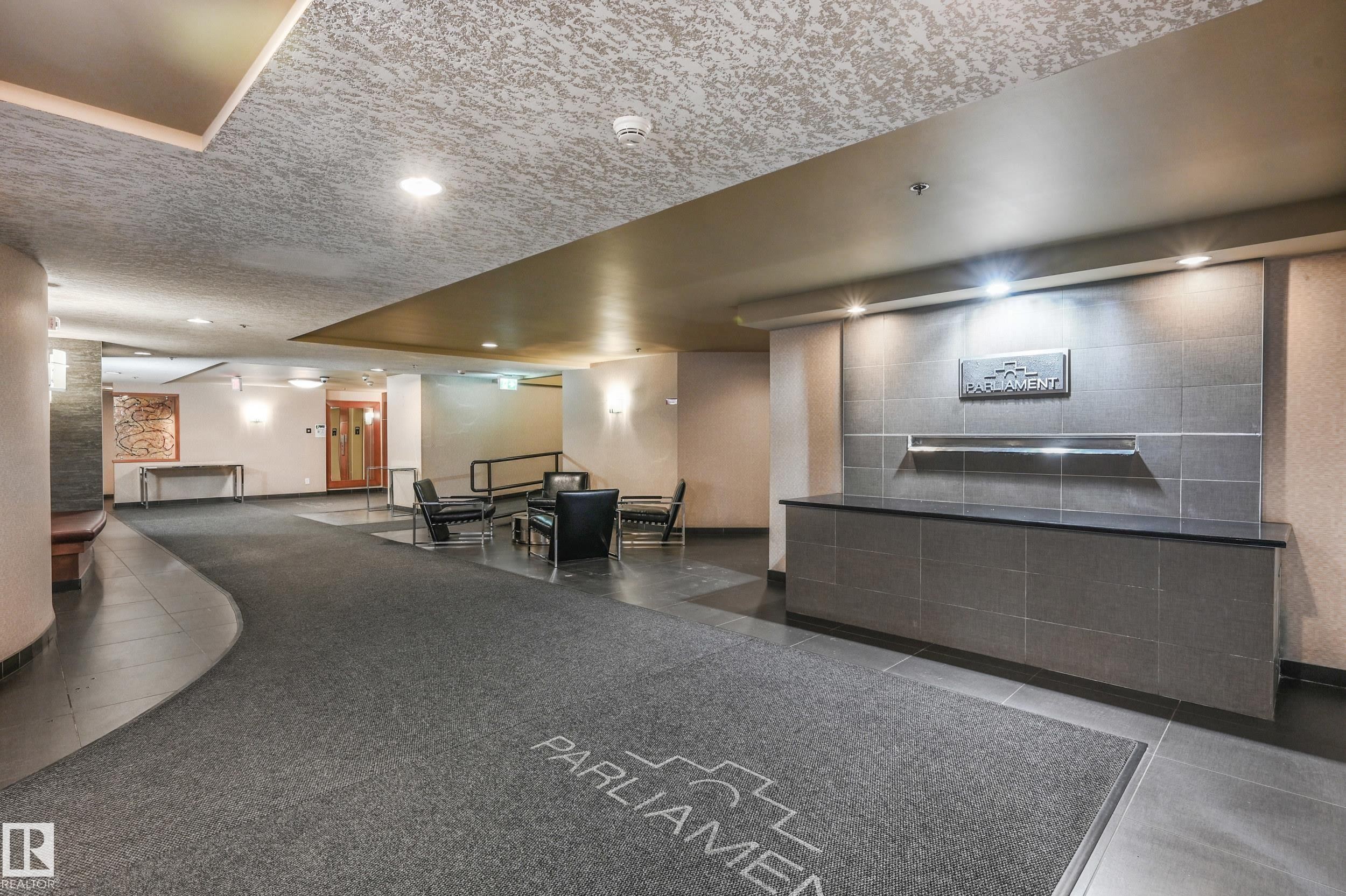 Building lobby featuring a reception desk with a dark countertop and tiled facade, contemporary wall sconces, recessed ceiling lighting, and a combination of carpet and hard-surface flooring - #1407 9939 109 St Nw, Edmonton, AB