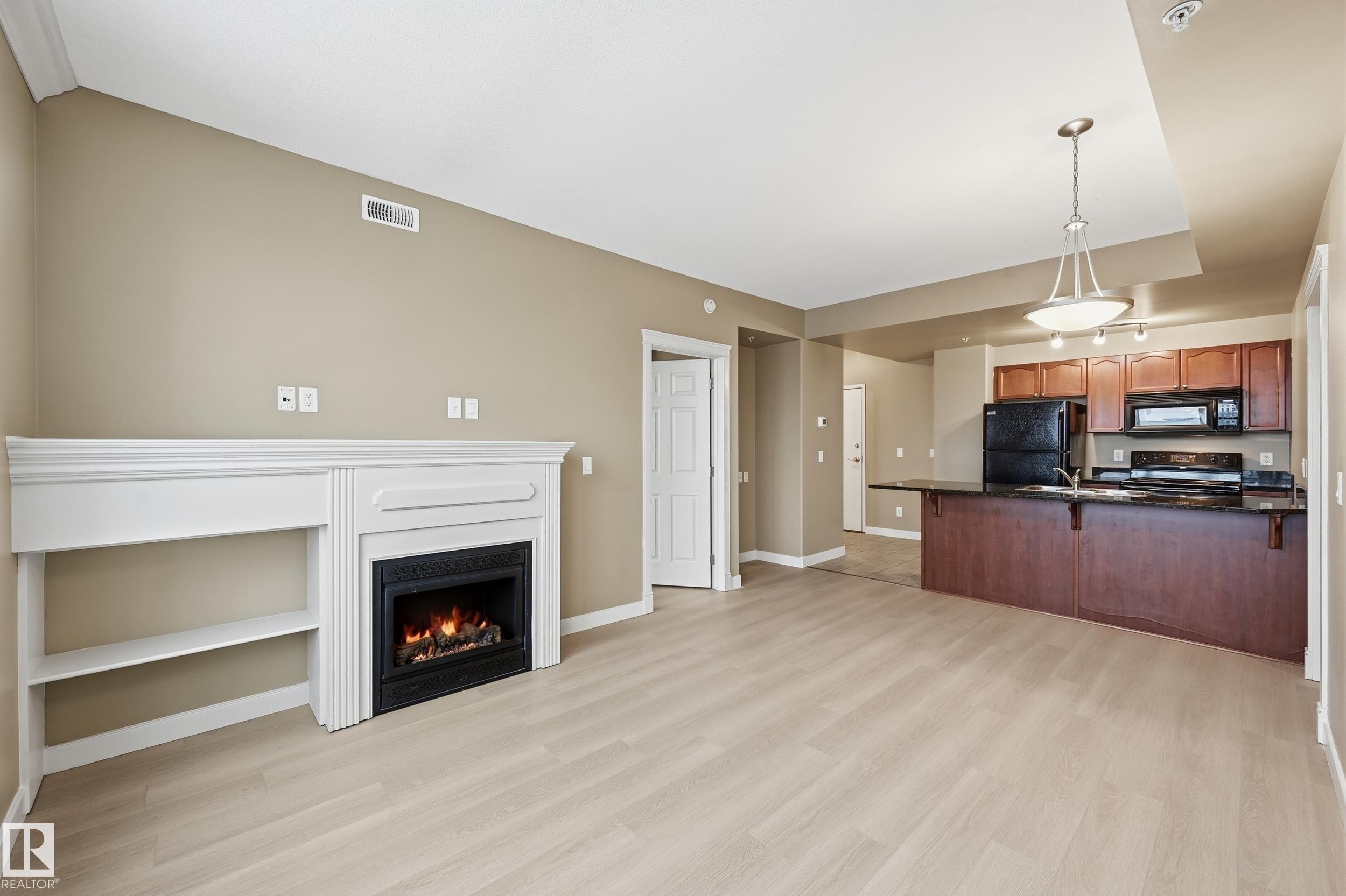 Open-concept living area featuring light wood-finish flooring, a built-in electric fireplace with white mantel and shelving, and a kitchen with dark cabinetry, black appliances, and a peninsula with dark countertops - #1407 9939 109 St Nw, Edmonton, AB