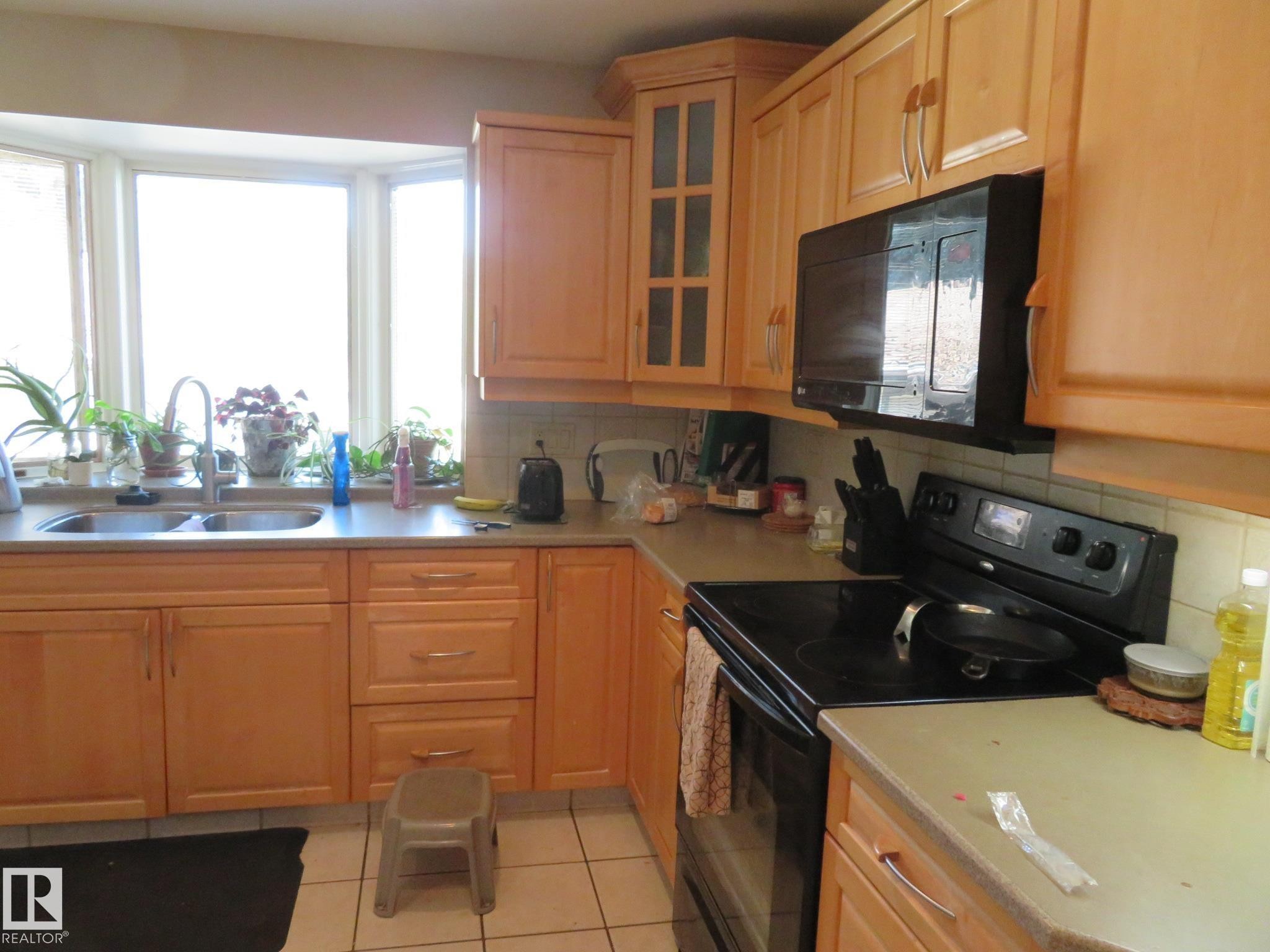 Kitchen featuring light wood cabinetry, a bay window with a double basin stainless steel sink, light-toned countertops, and an over-the-range microwave - 10720 57 Avenue Nw, Edmonton, AB - Indoor Photo Showing Kitchen With Double Sink