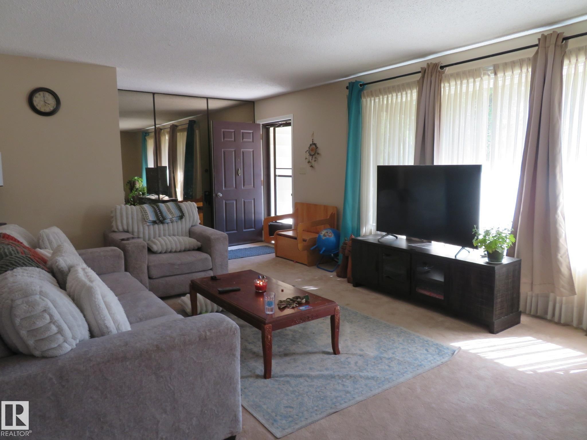 Living room featuring neutral-toned walls, light carpet flooring, large windows with vertical blinds and curtain rods, and a dark wood-finish media console - 10720 57 Avenue Nw, Edmonton, AB - Indoor Photo Showing Living Room