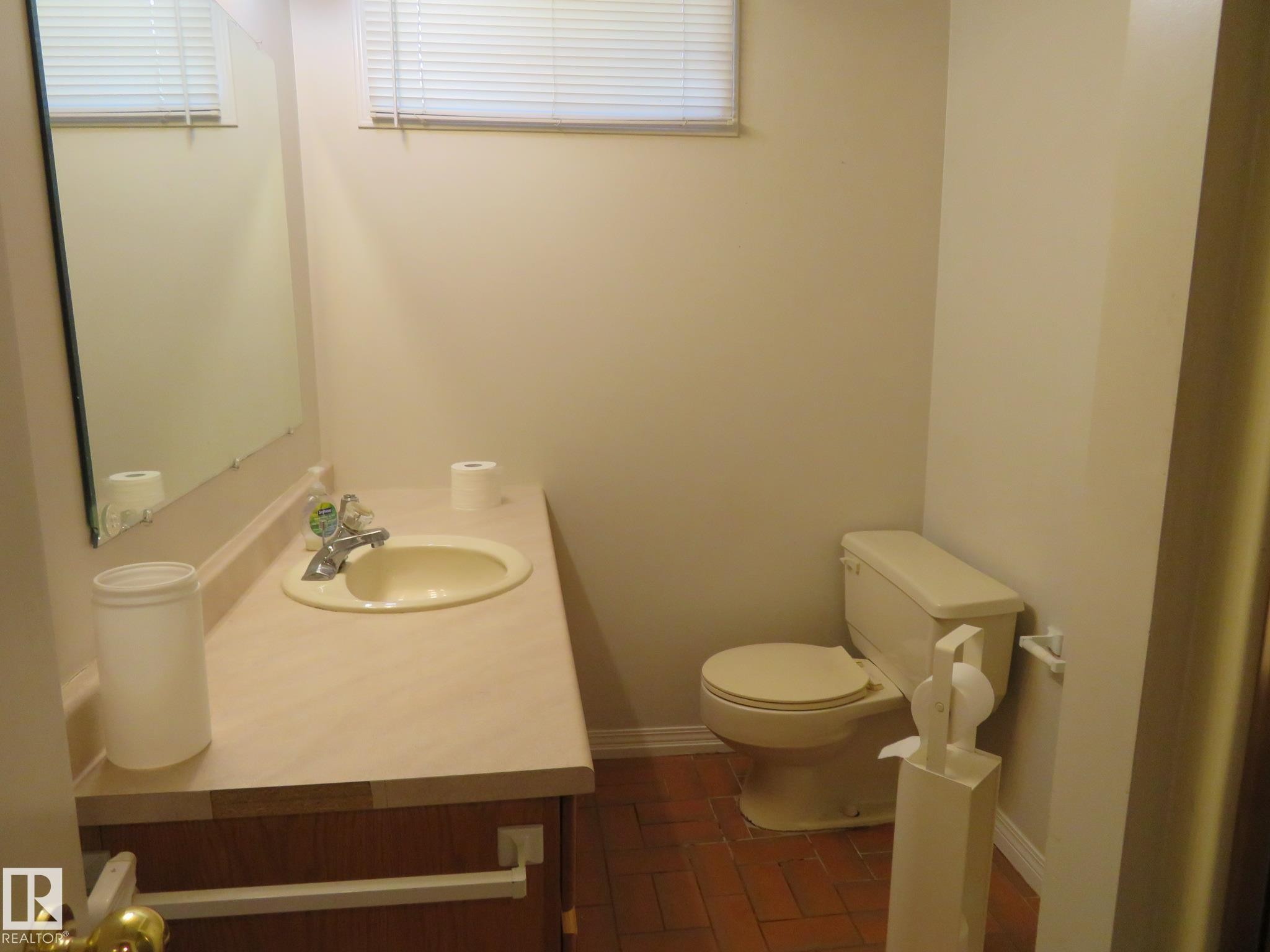 Bathroom featuring a vanity with an integrated sink, a rectangular mirror, a window with blinds, and brick-patterned flooring - 10720 57 Avenue Nw, Edmonton, AB - Indoor Photo Showing Bathroom