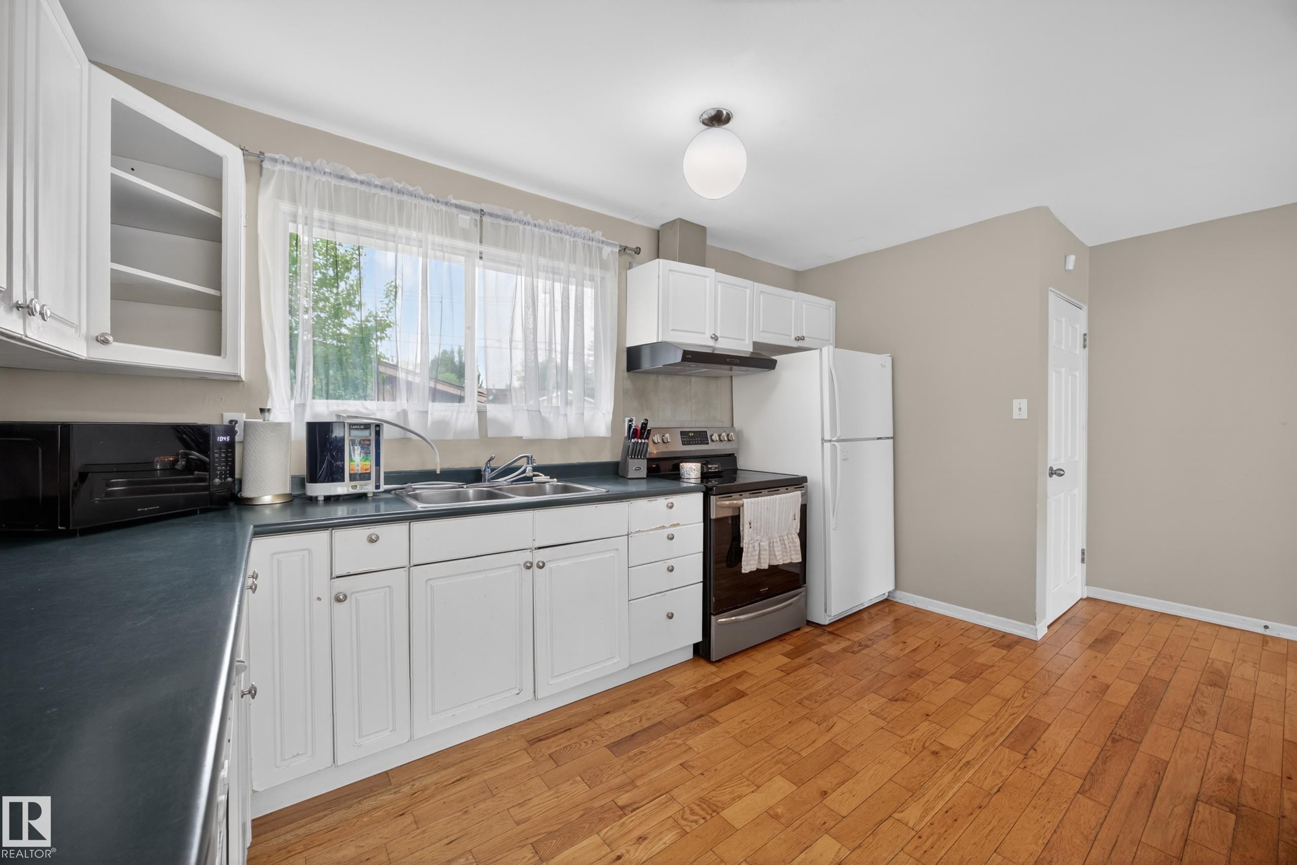 Kitchen featuring wood-finish flooring, white cabinetry, dark countertops, a double basin sink, and a large window - 5216 104A Street, Edmonton, AB - Indoor Photo Showing Kitchen With Double Sink