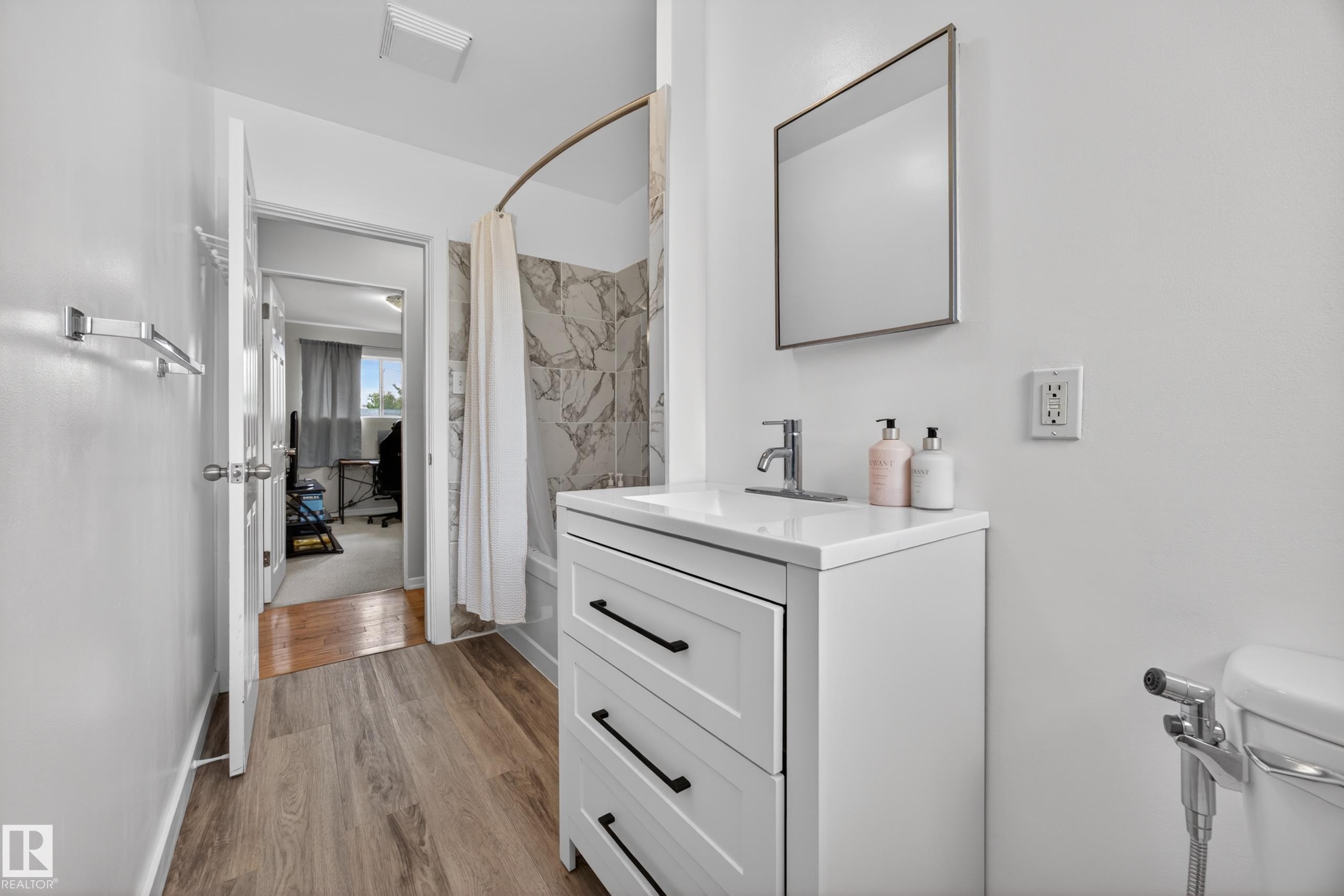 Bathroom featuring wood-finish flooring, a white vanity with black hardware, a rectangular sink, and a framed mirror - 5216 104A Street, Edmonton, AB - Indoor