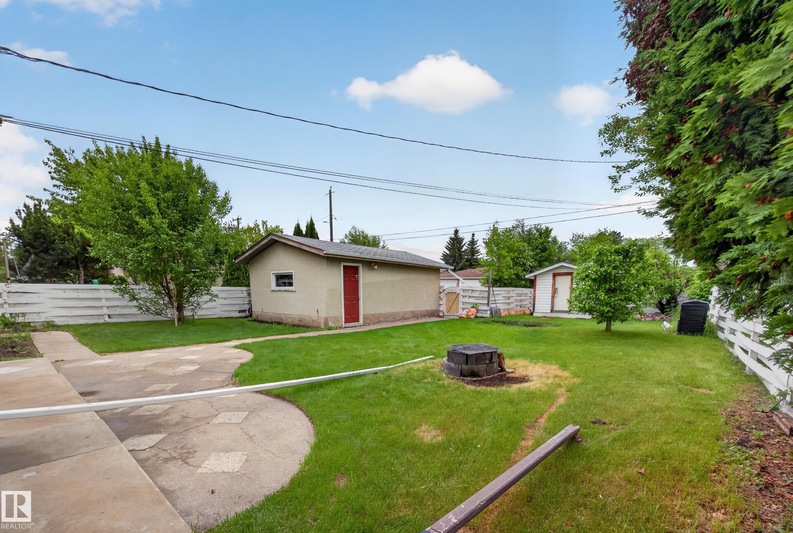 Spacious backyard featuring a stucco detached garage with a red door, a storage shed, a manicured lawn, and a white picket fence - 5216 104A Street, Edmonton, AB - Outdoor