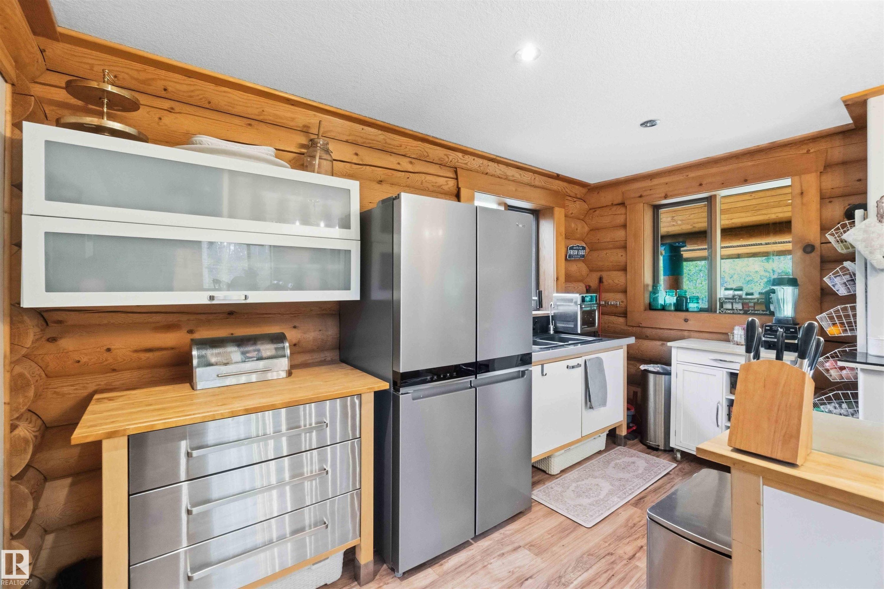 Kitchen featuring log wall construction, wood-finish flooring, a stainless steel refrigerator, white cabinetry, and a window with a wood frame - #17 A 15031 Twp Road 462, Rural Wetaskiwin County, AB - Indoor Photo Showing Kitchen