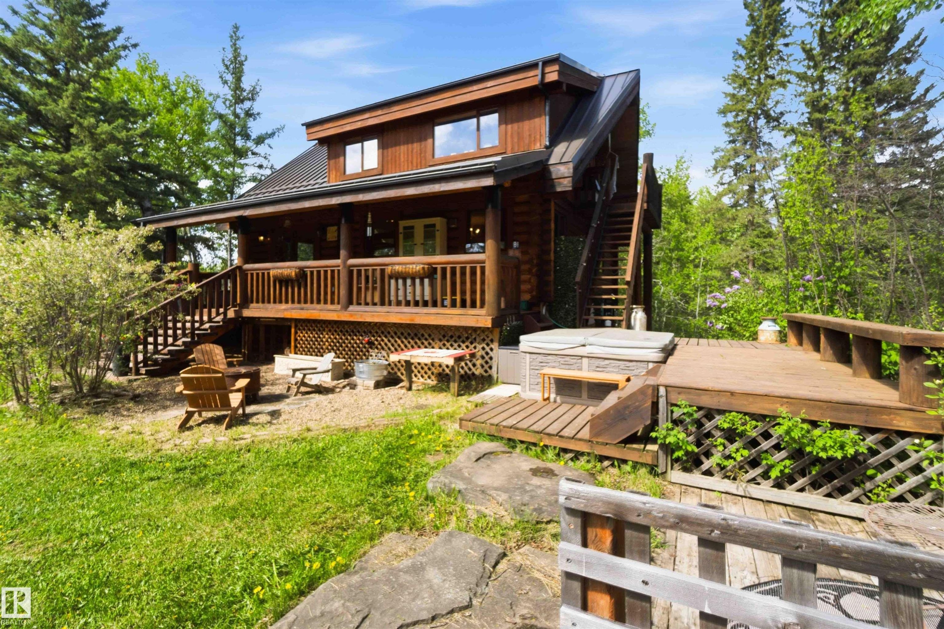 Wood-sided dwelling with a dark metal roof, featuring a covered porch, an exterior staircase, a ground-level patio, and an adjacent wood deck with an integrated hot tub - #17 A 15031 Twp Road 462, Rural Wetaskiwin County, AB - Outdoor With Deck Patio Veranda