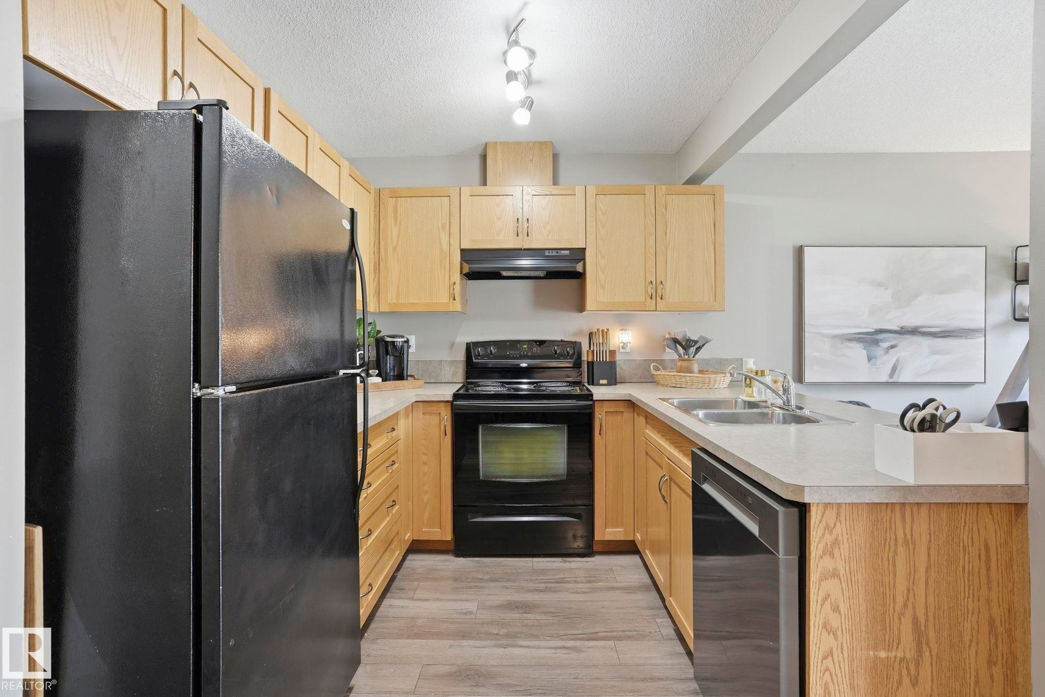 U-shaped kitchen featuring light wood cabinetry, stone-finish countertops, black appliances, and wood-finish flooring - 164 150 Edwards Drive, Edmonton, AB - Indoor Photo Showing Kitchen With Double Sink