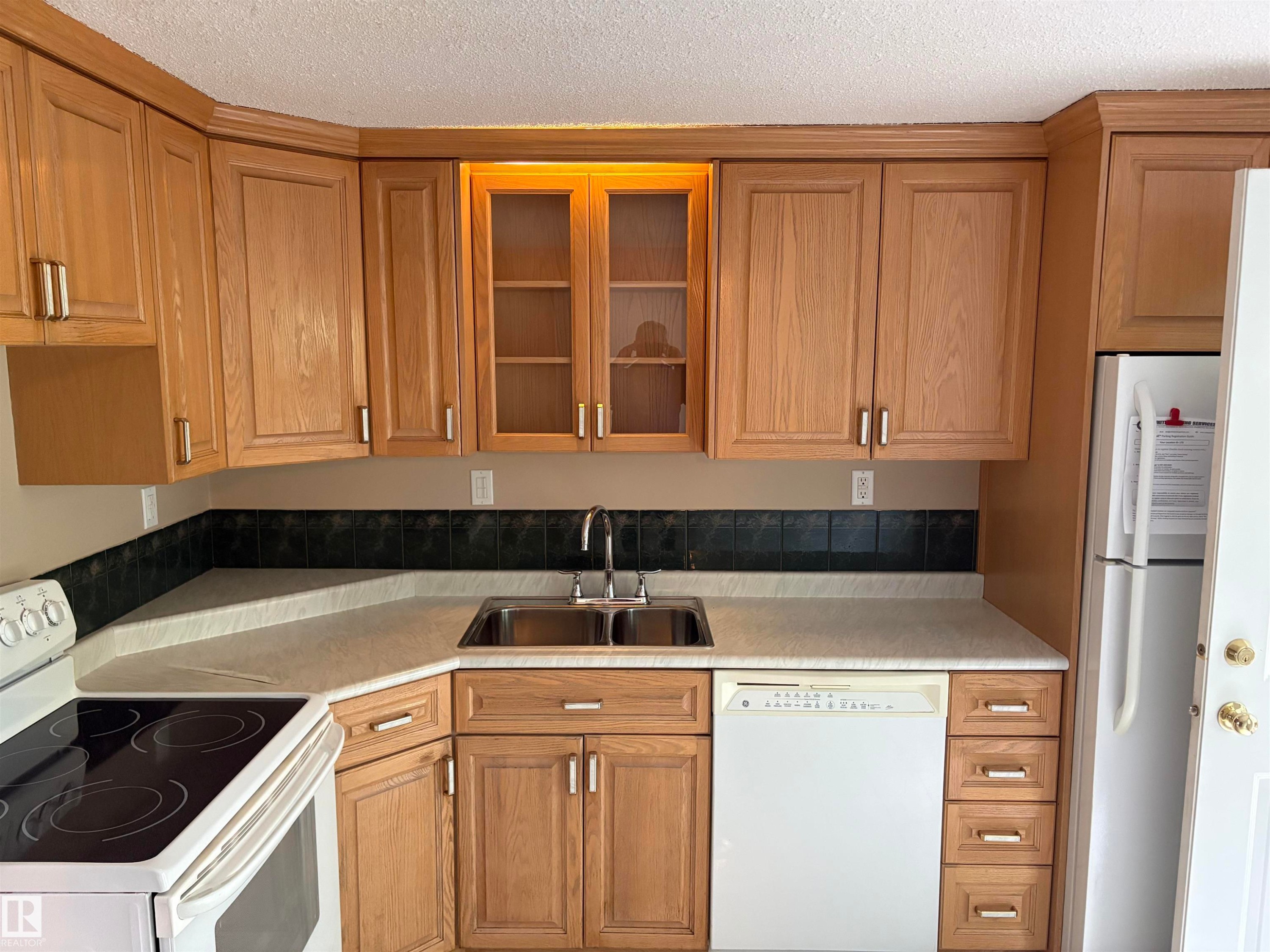 Kitchen featuring wood-finish cabinetry with modern hardware, a double basin stainless steel sink, light-toned countertops, and a dark tile backsplash - 167 Ridgewood, St. Albert, AB - Indoor Photo Showing Kitchen With Double Sink