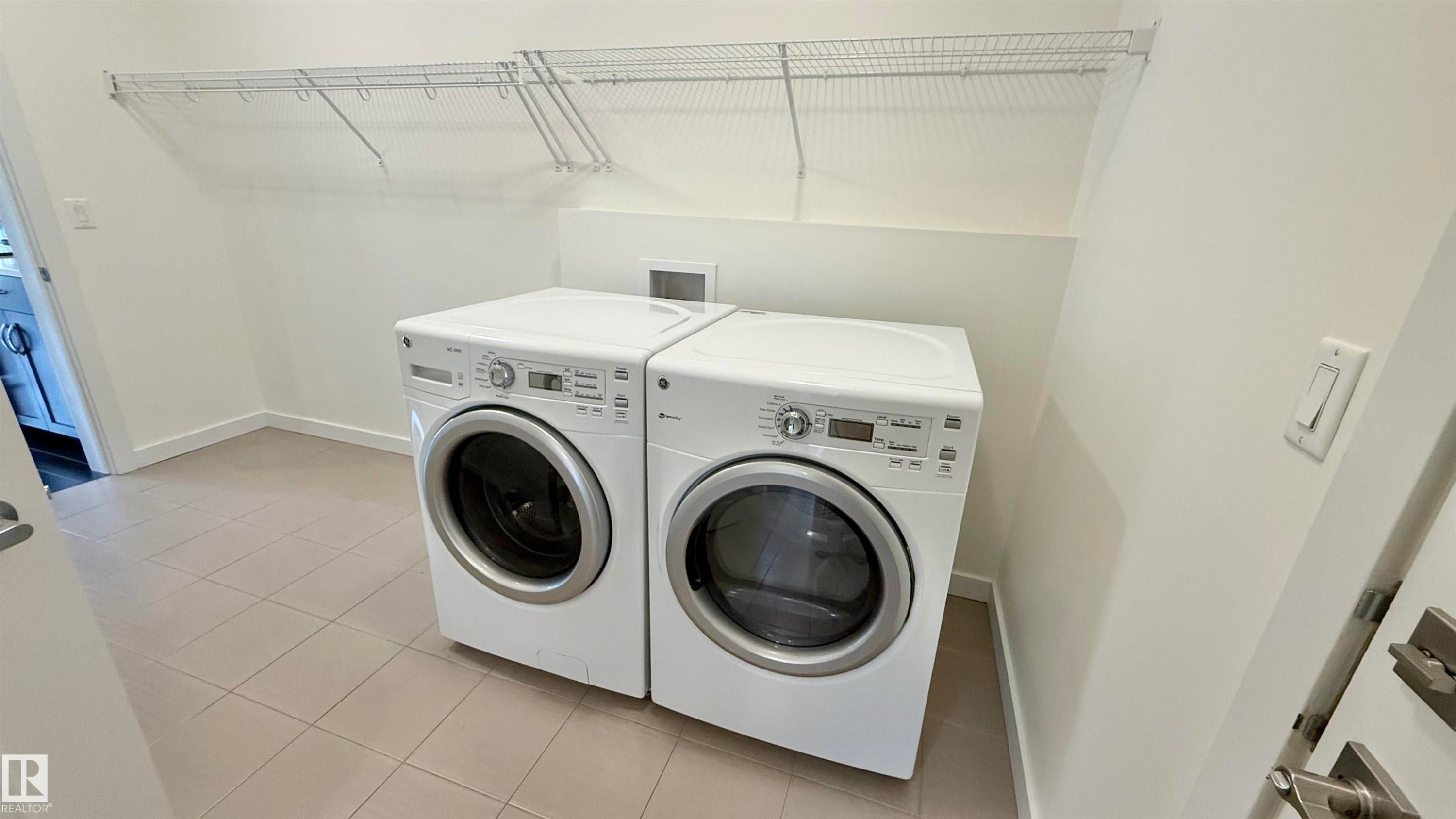 Dedicated laundry area featuring front-loading appliances, neutral-toned floor tile, white perimeter baseboards, and overhead wire shelving - 3962 Claxton Loop, Edmonton, AB - Indoor Photo Showing Laundry Room